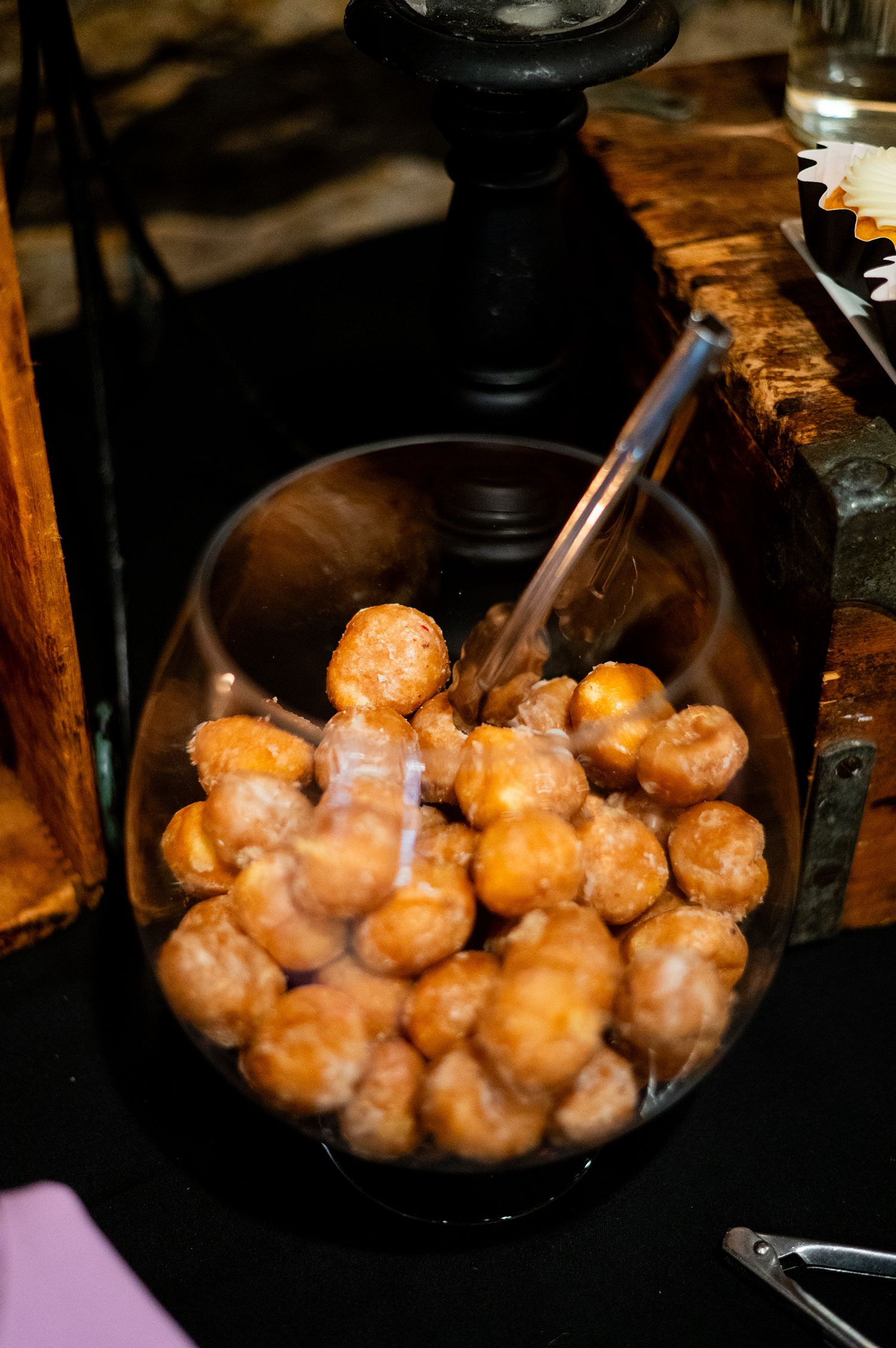 Golden glazed donut holes served in a silver bowl with tongs at a catering event.