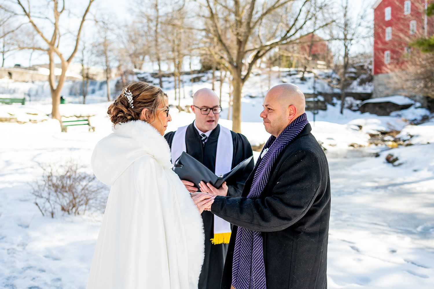 Winter wedding ceremony taking place outdoors in snowy park setting with white cape and formal attire.