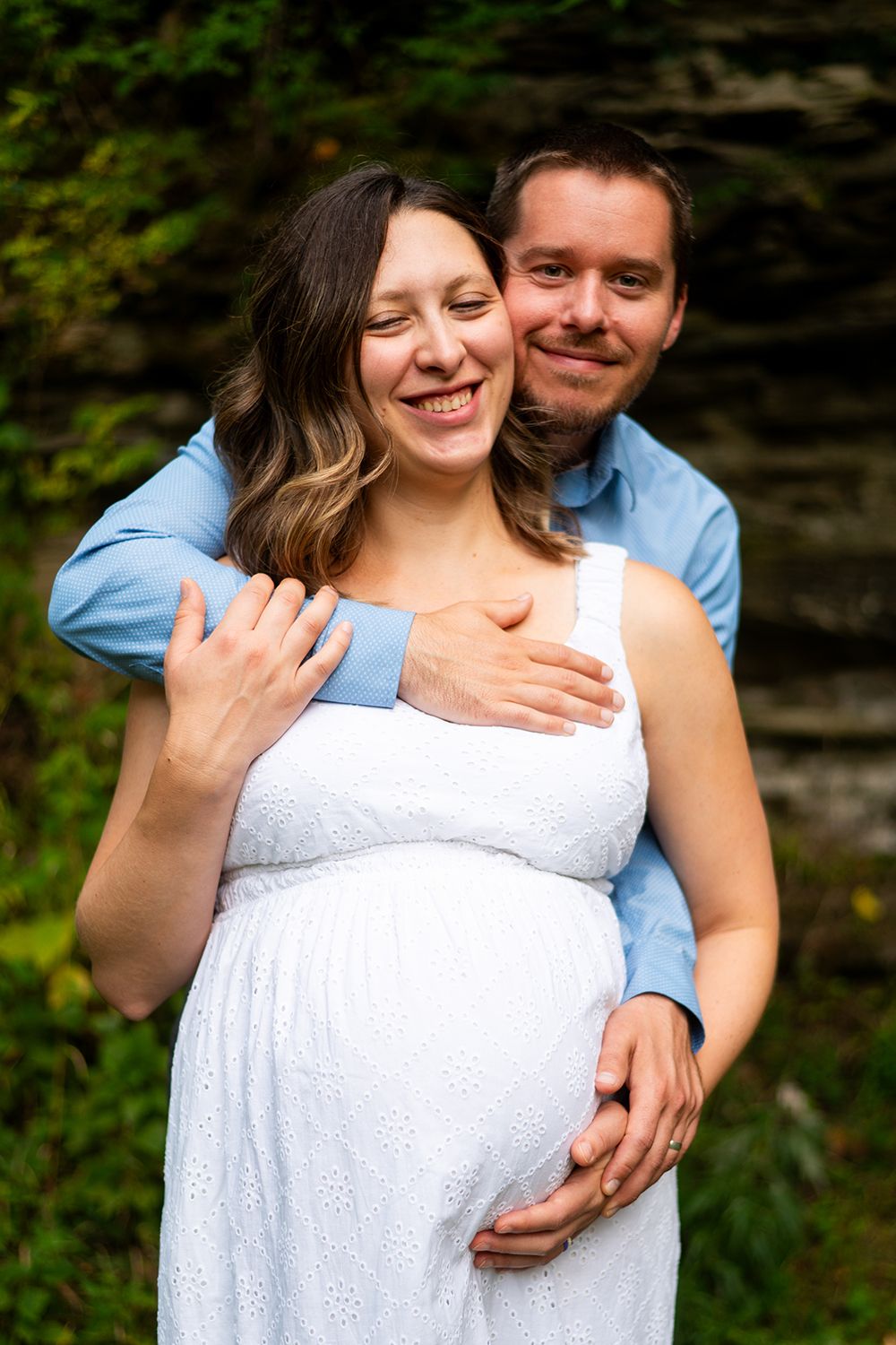 A couple embraces outdoors in front of a natural rock wall, wearing a white dress and blue shirt.