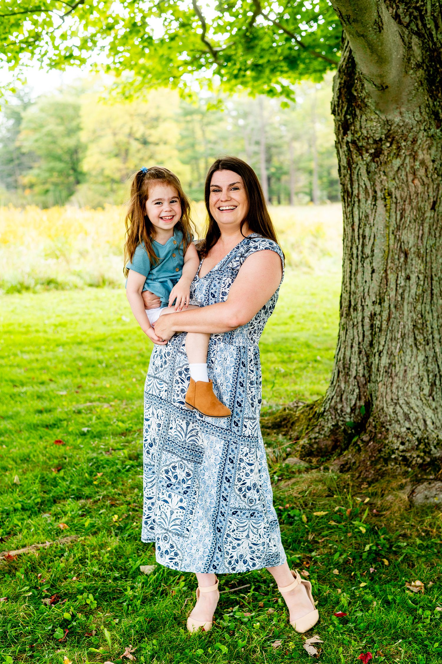 A smiling woman in a blue floral dress holds a young girl outdoors near a large tree on a green lawn.