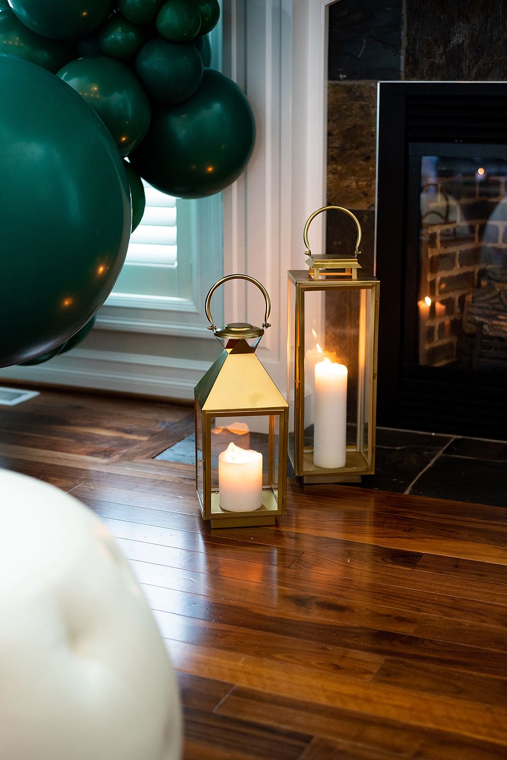 Gold lanterns with glowing candles sit on hardwood flooring beside green party balloons and a fireplace.