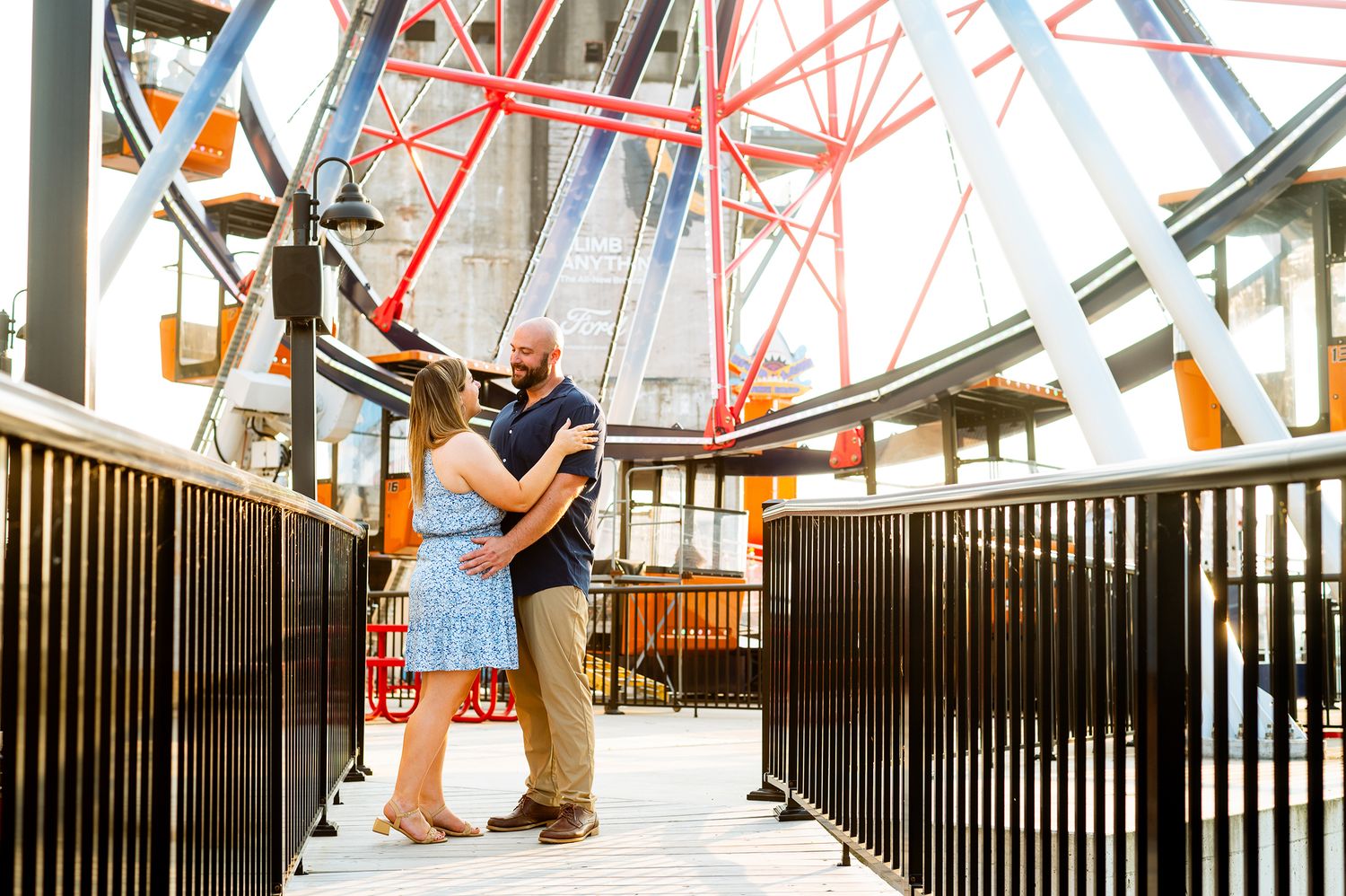 Couple embracing on a bridge near a colorful Ferris wheel at an amusement park during an engagement session.