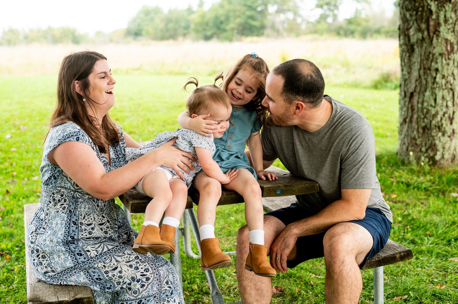 Family of four enjoying a sunny day outdoors on a picnic bench, with parents smiling at their two young children.