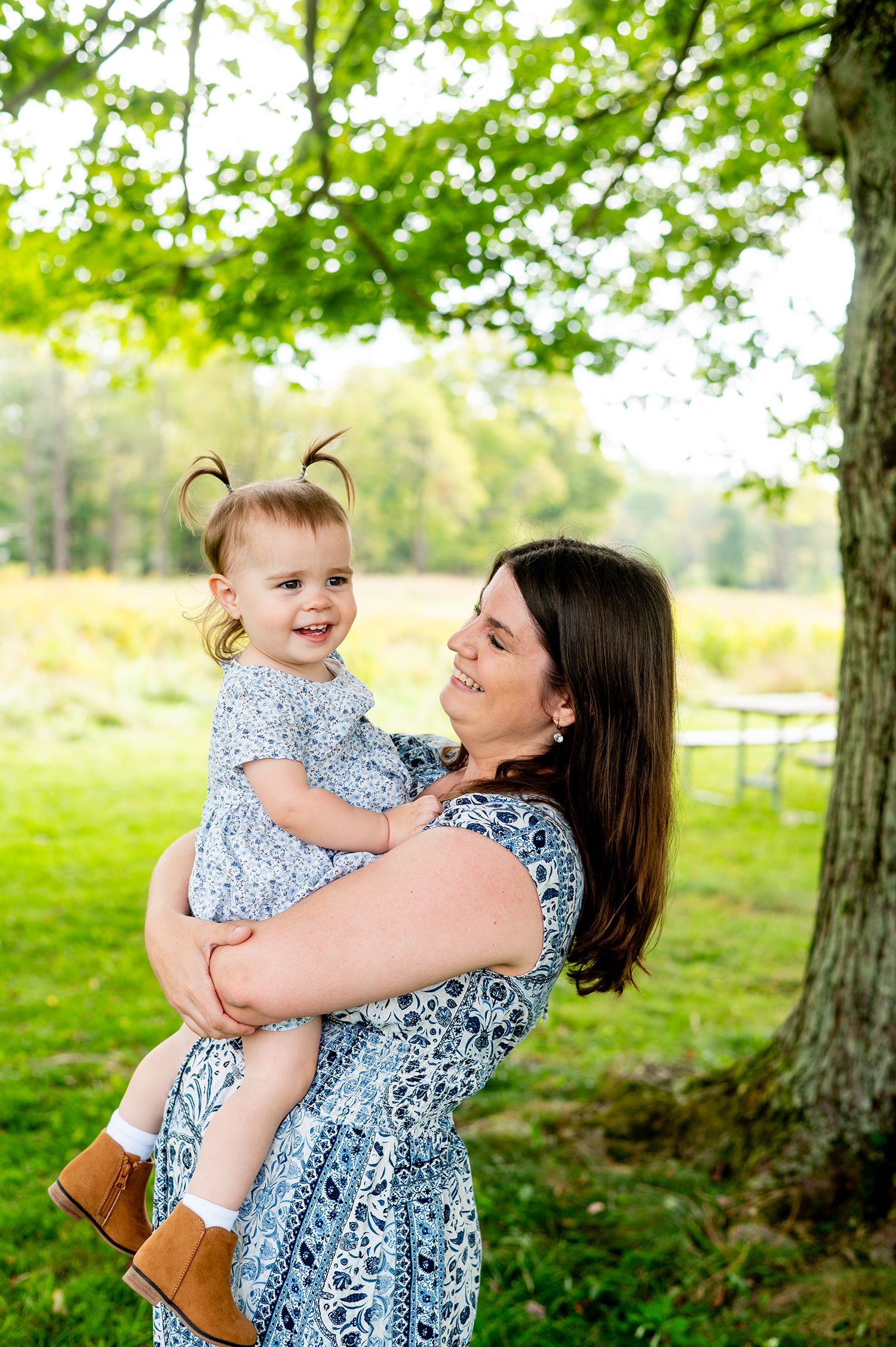 A mother in a blue floral dress holds her smiling toddler girl outdoors under a green tree.