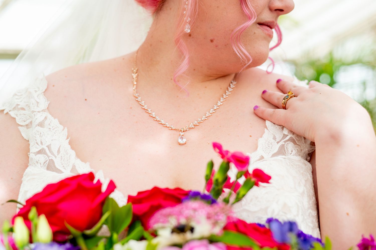 Bride in white lace dress holding colorful bouquet with pink hair, pearl necklace, and gold ring on wedding day.