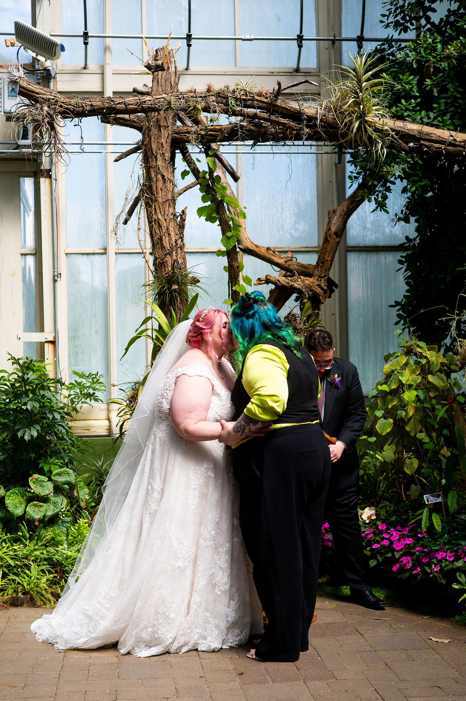 Two brides sharing a kiss at a greenhouse wedding ceremony, one in white lace gown with pink hair, other in yellow jacket with teal hair.