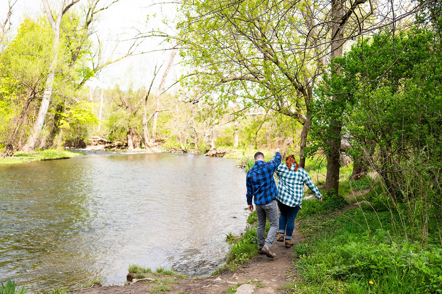 Two people in casual clothing standing on a riverbank path surrounded by green spring foliage and flowing water.