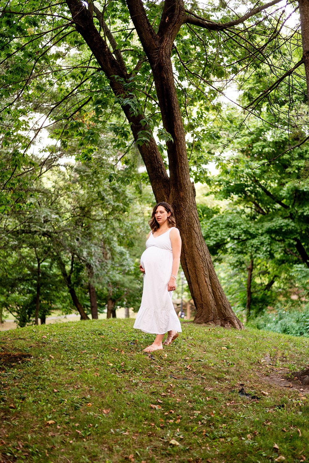 A person in a flowing white dress stands gracefully under a large tree in a lush green garden setting.