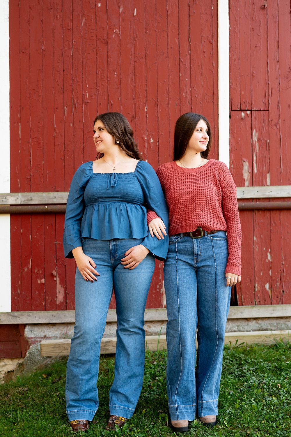 Two women in blue jeans stand against a rustic red barn wall wearing blue and red sweaters.