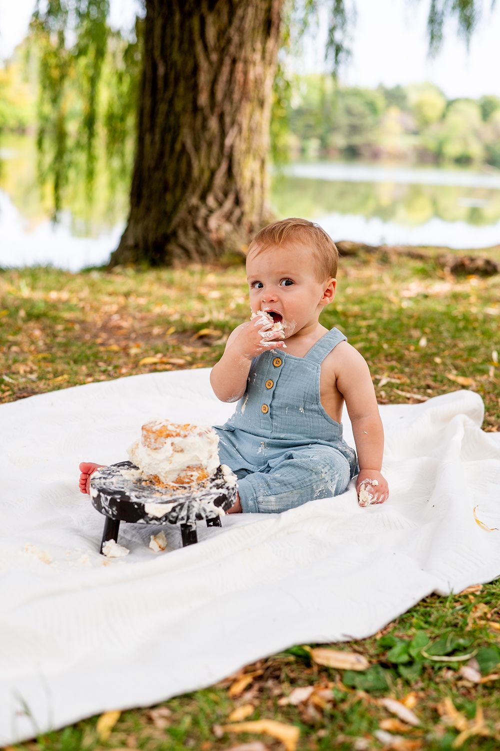 Small child in denim overalls enjoying cake while sitting on a white blanket under a willow tree by a lake.