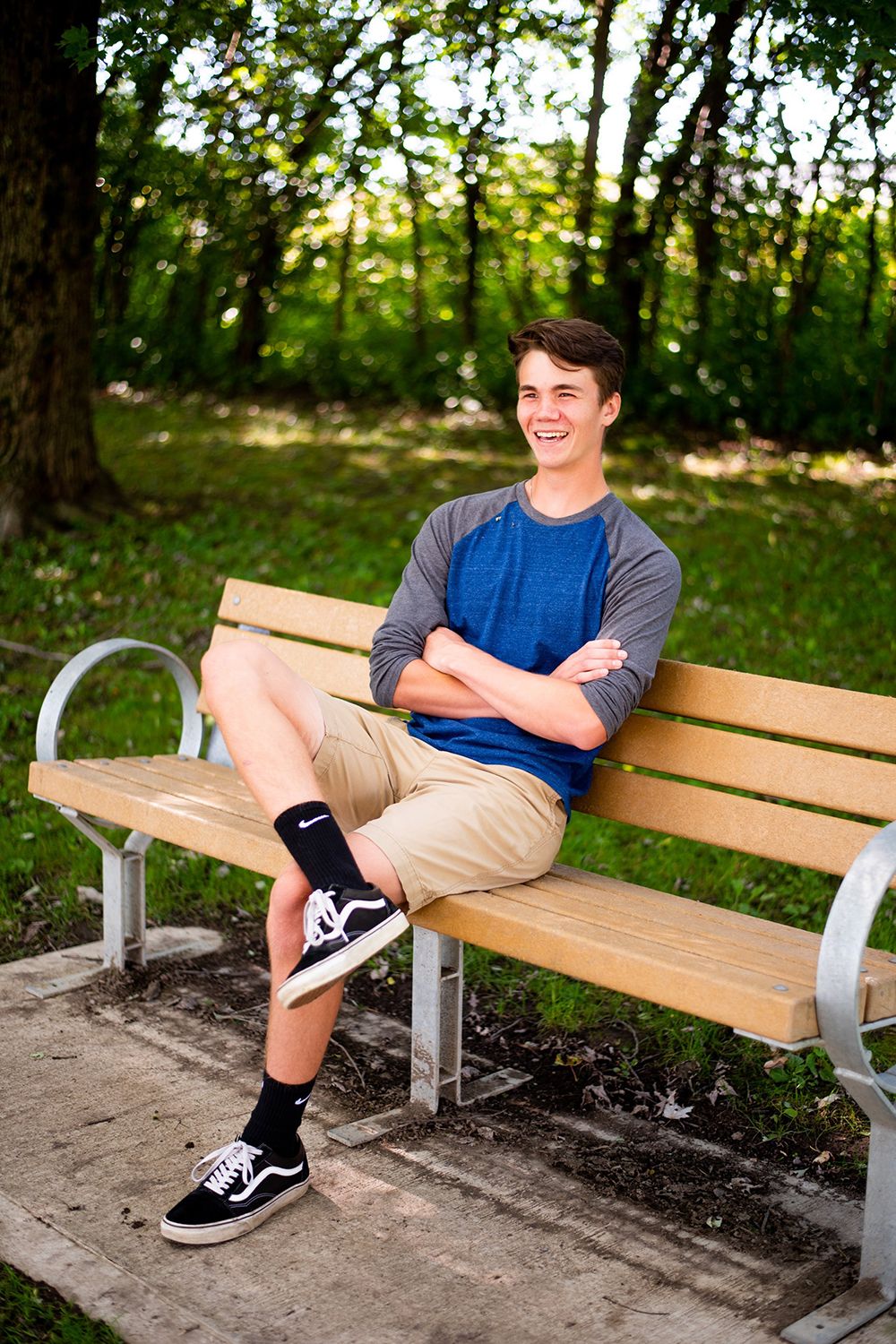 A casual portrait of someone relaxing on a wooden park bench wearing a blue raglan shirt and khaki shorts.