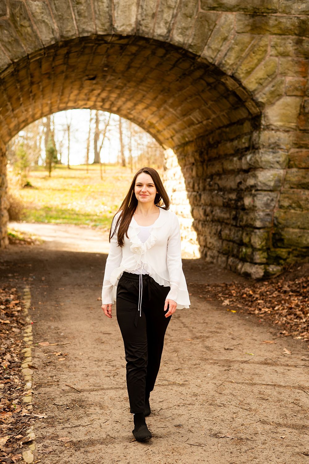 A person walks beneath a stone archway tunnel on a dirt path surrounded by autumn leaves and trees in natural lighting.