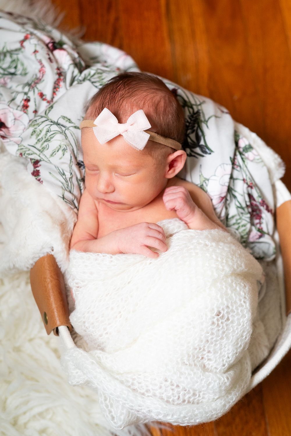 Peaceful sleeping newborn swaddled in white blanket with delicate white bow headband laying on floral bedding.