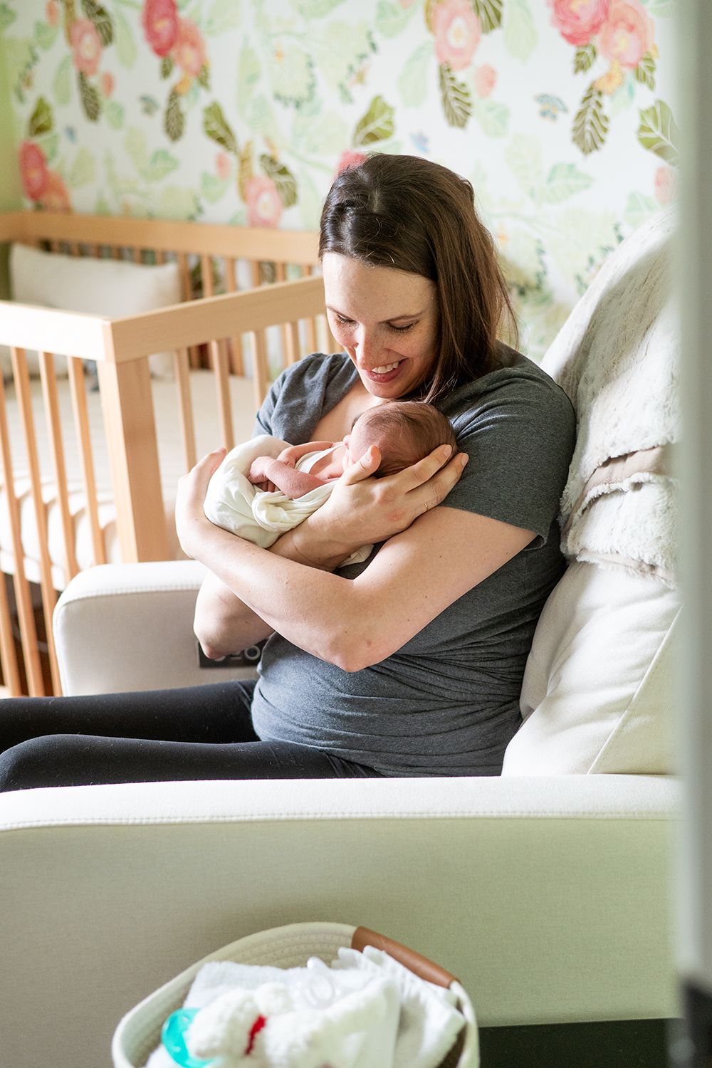 Mother tenderly holding newborn baby in nursery with floral wallpaper and wooden crib.