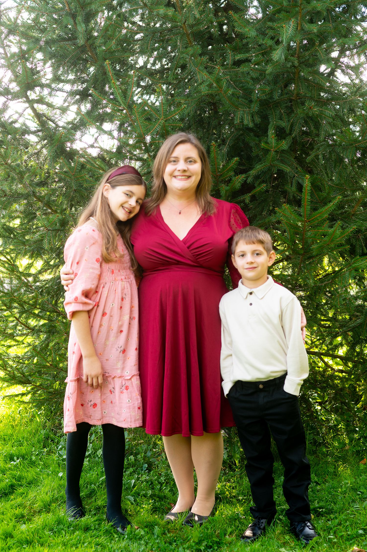 A woman in a red dress poses outdoors with a girl in pink and a boy in white shirt, standing before evergreen trees.