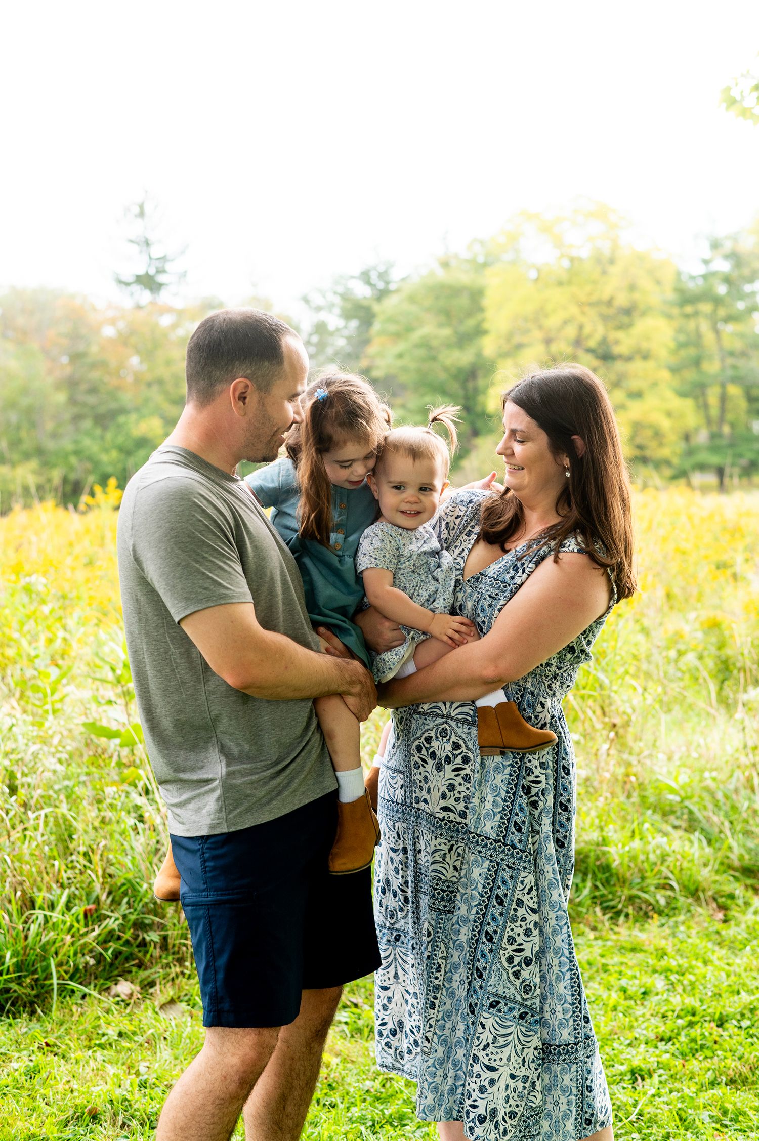 A happy family of four enjoys a sunny outdoor moment in a green meadow, with parents holding two young daughters.