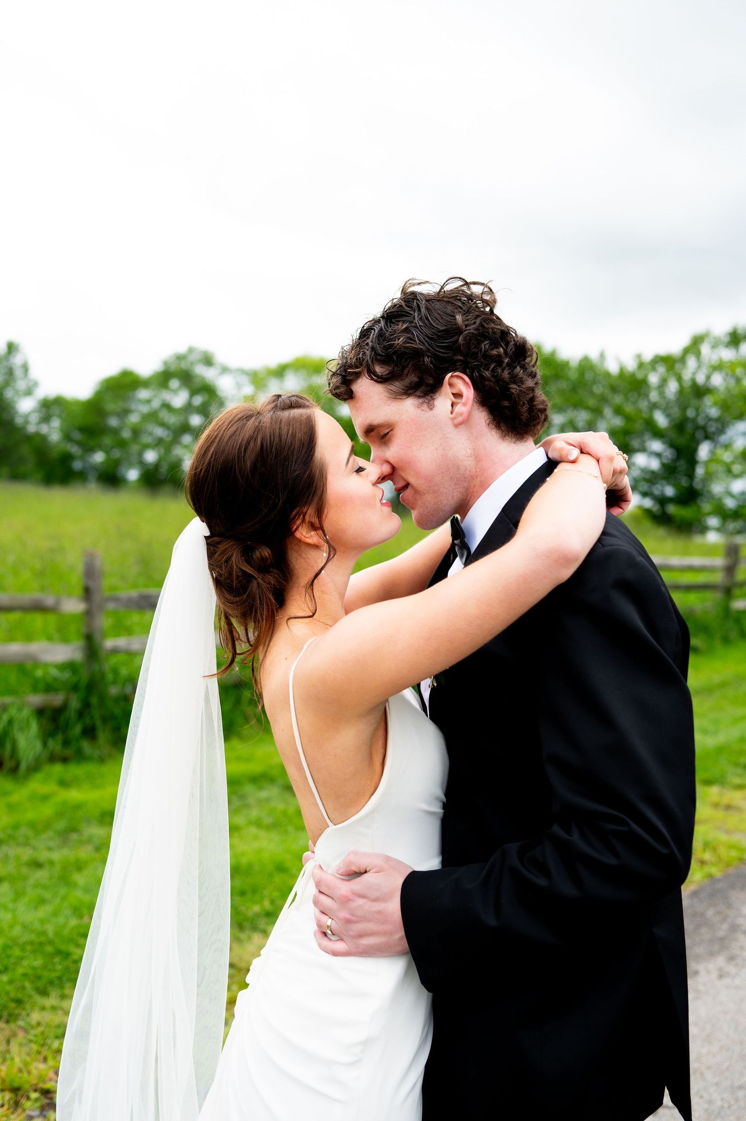 A romantic wedding couple shares a tender kiss outdoors with green trees and cloudy skies in the background.