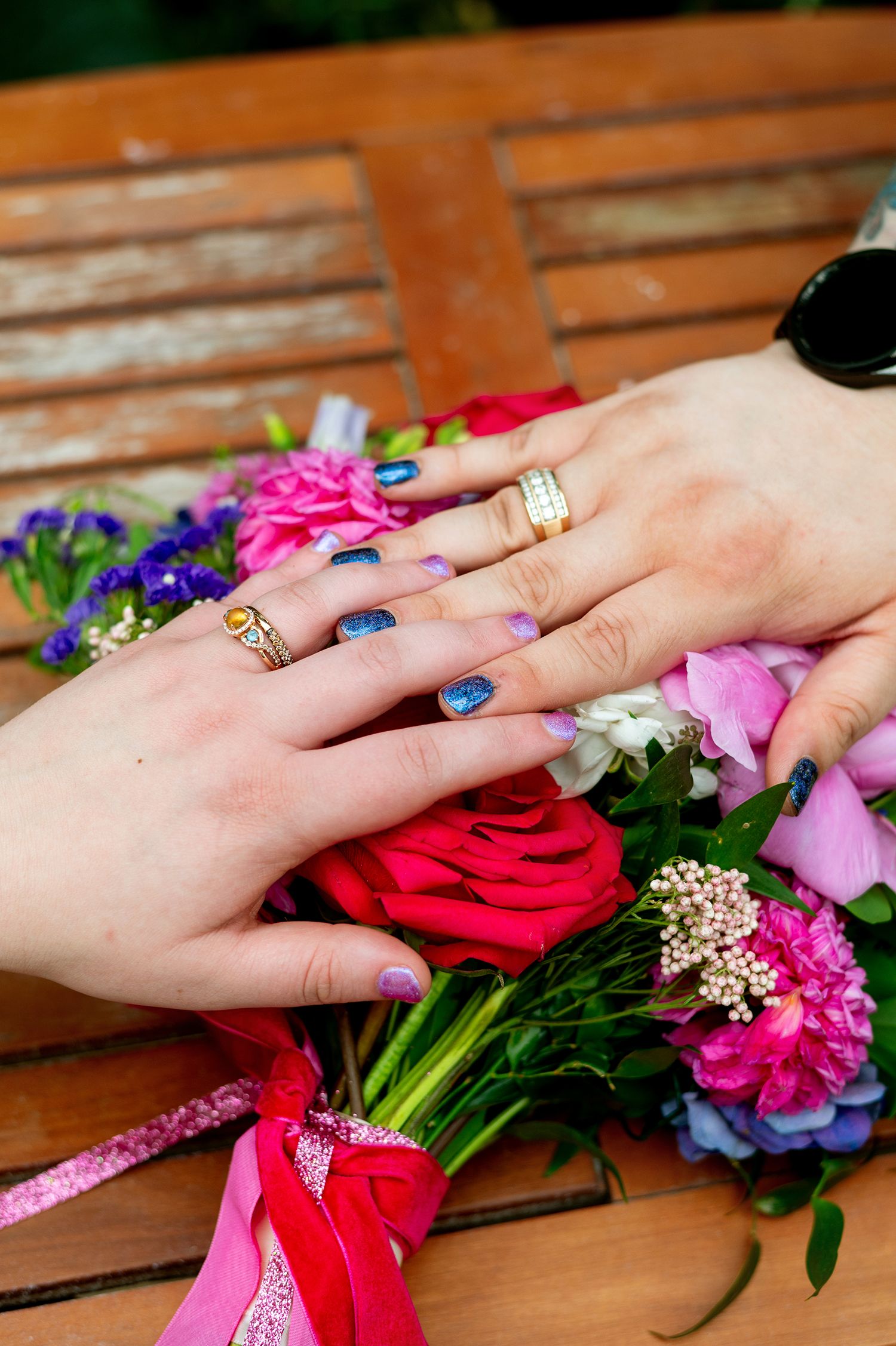 Two hands with rings and blue nail polish rest on a colorful bridal bouquet of roses and wildflowers on a wooden bench.