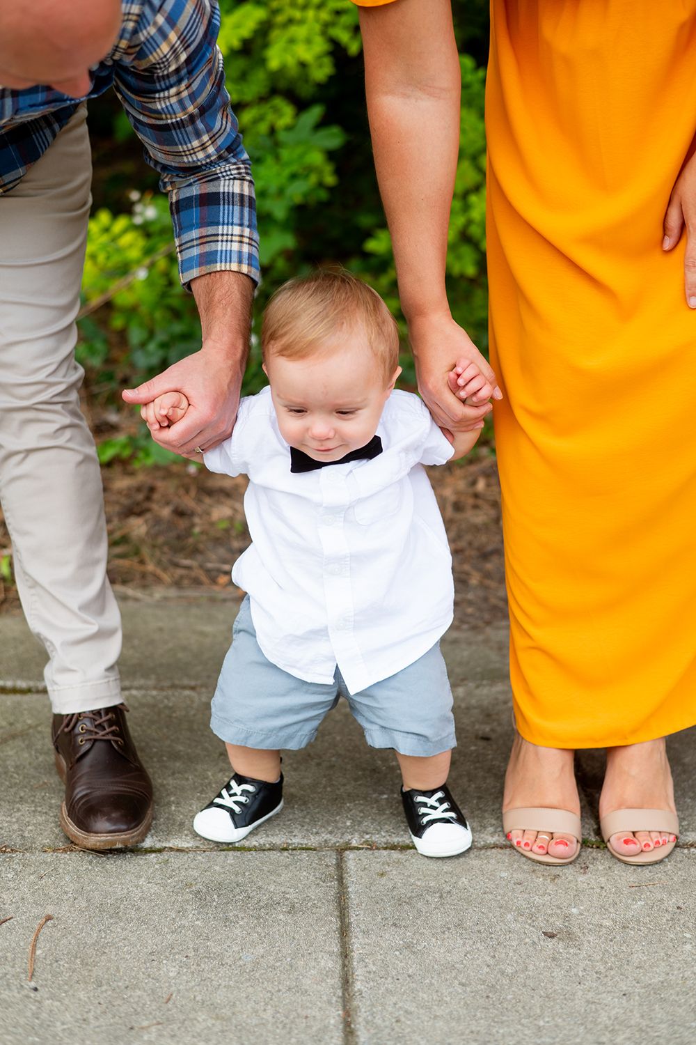 A small child in white shirt and shorts takes first steps while holding hands with adults in colorful attire.