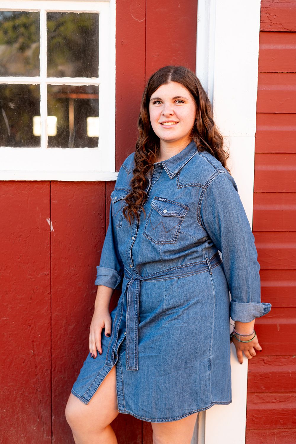 Woman in denim shirt dress poses outdoors against red building with white trim.