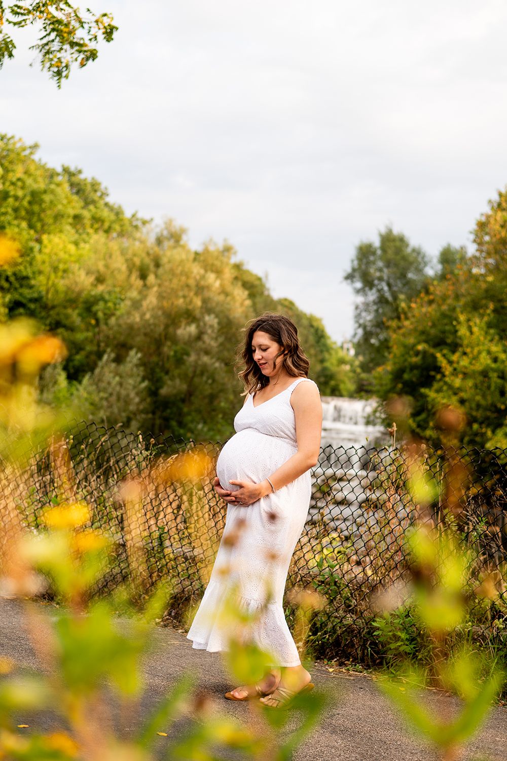 A maternity photo taken outdoors with autumn foliage and a waterfall in the background on a cloudy day.