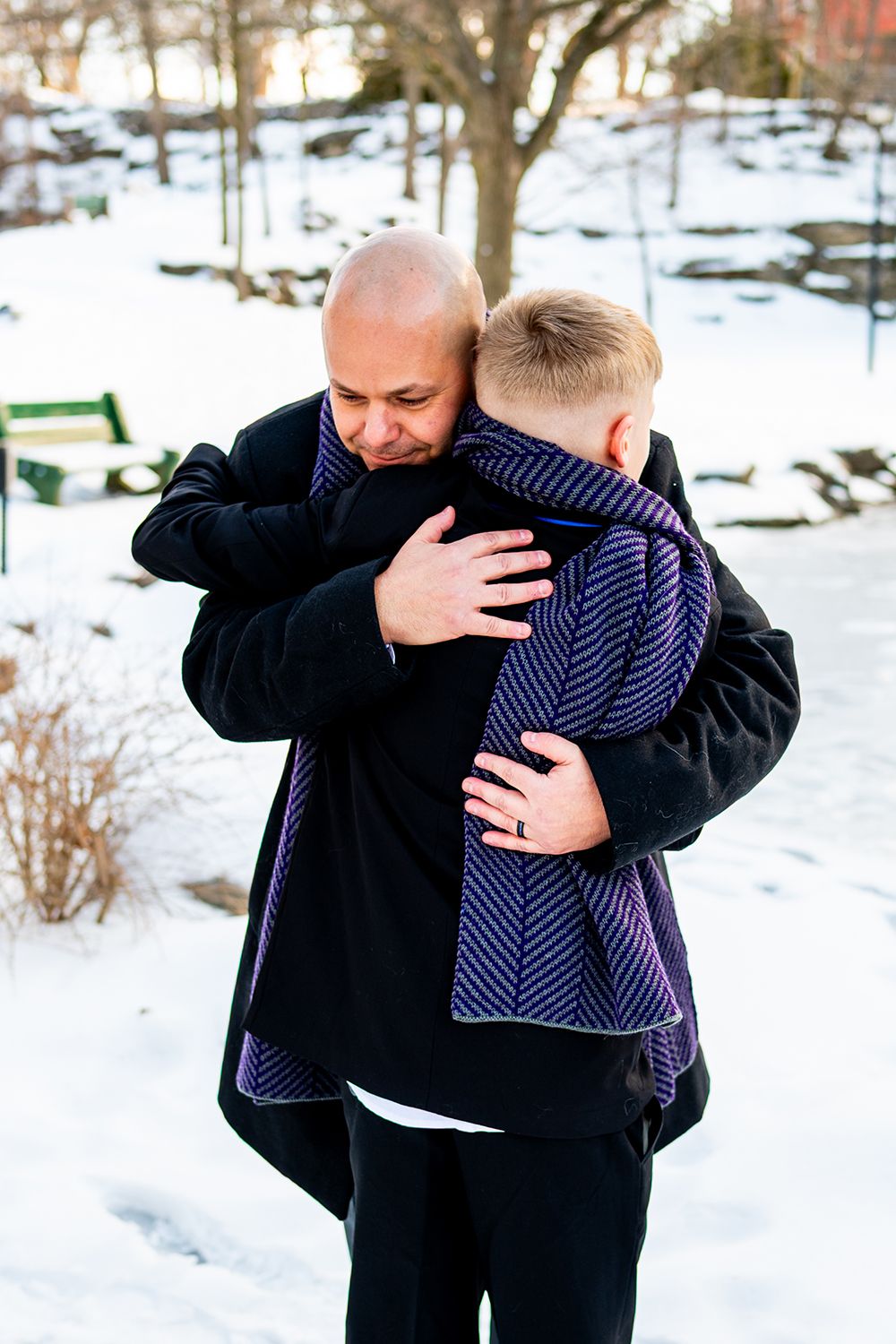 Two people sharing an emotional embrace in a snowy winter setting with trees and park benches in the background.