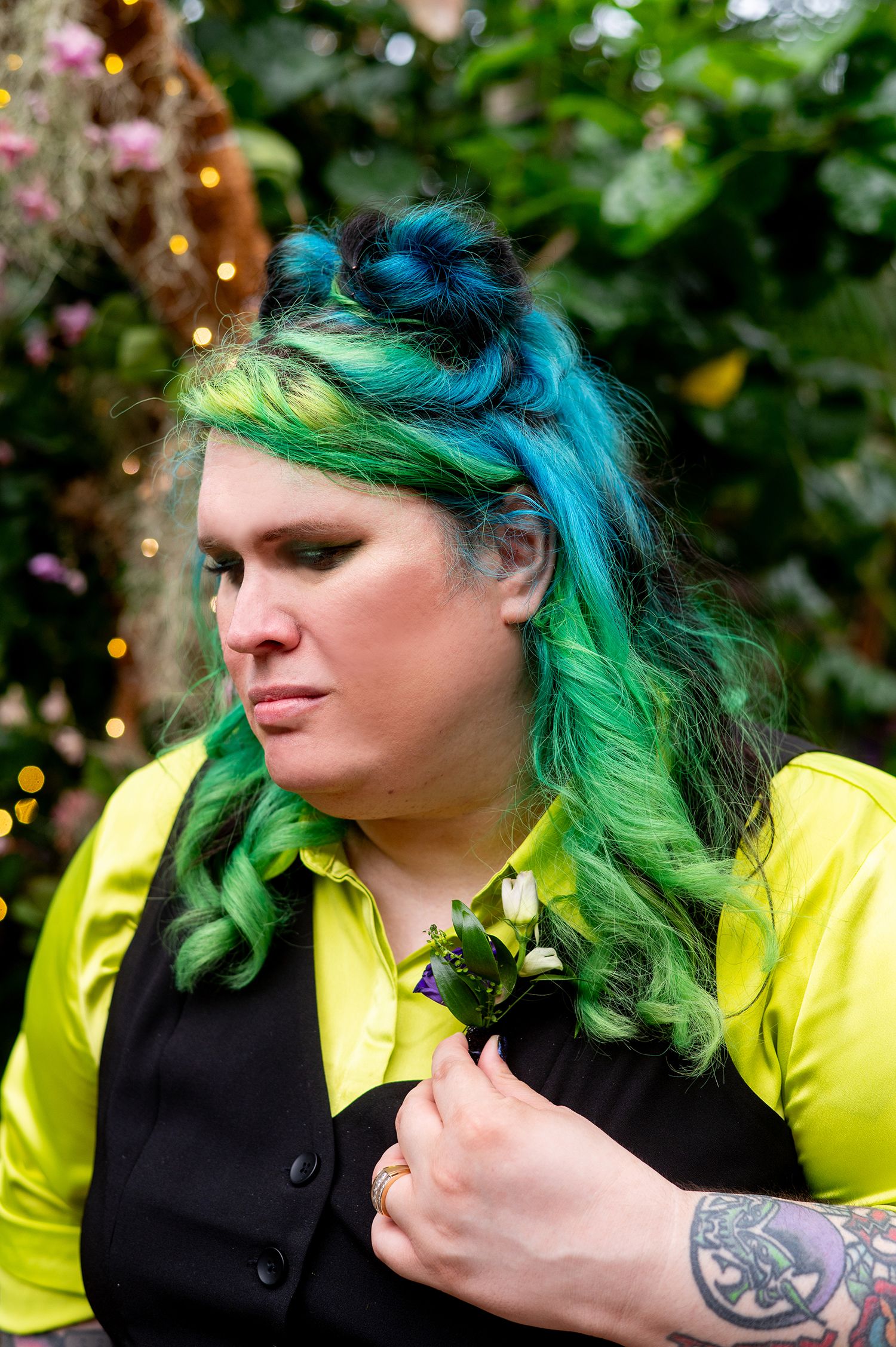 Person with vibrant green and teal hair wearing a yellow shirt and black vest with a floral boutonniere.