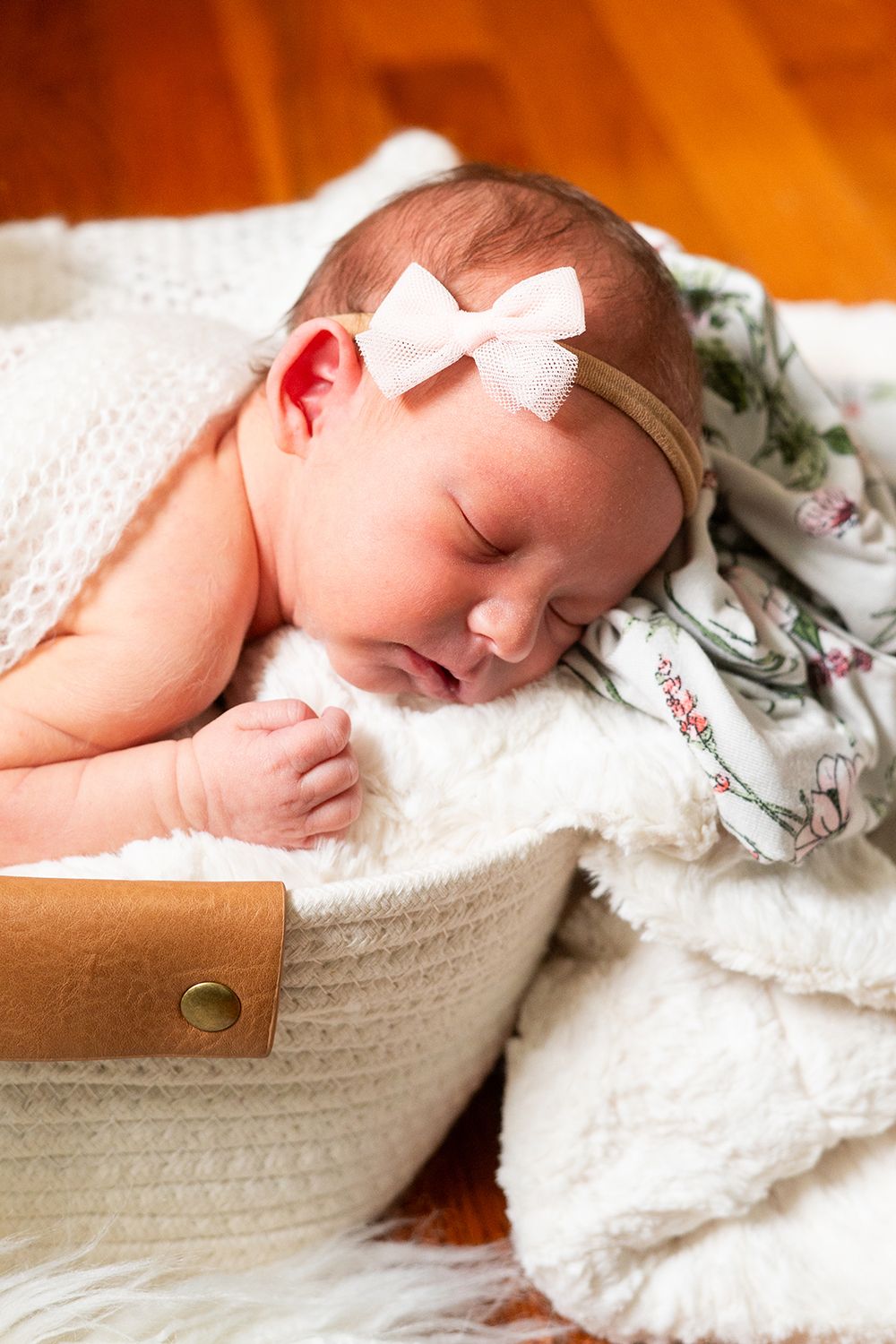 Sleeping baby wearing white bow headband rests peacefully in white basket with floral blanket and soft lighting.