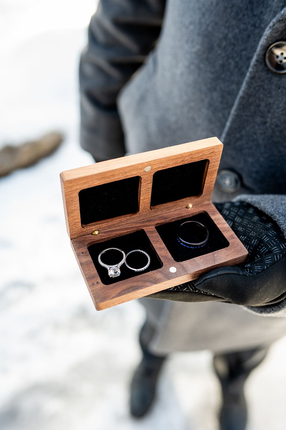 A wooden ring box with two wedding rings rests in the snow during an outdoor winter wedding ceremony.