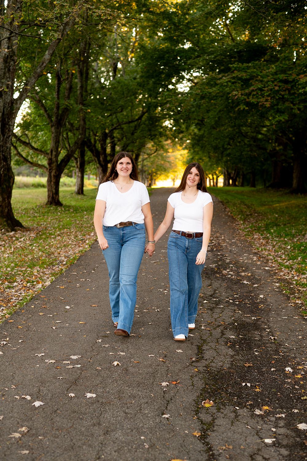 Two people in white tops and jeans walk together holding hands on a tree-lined path during golden hour.