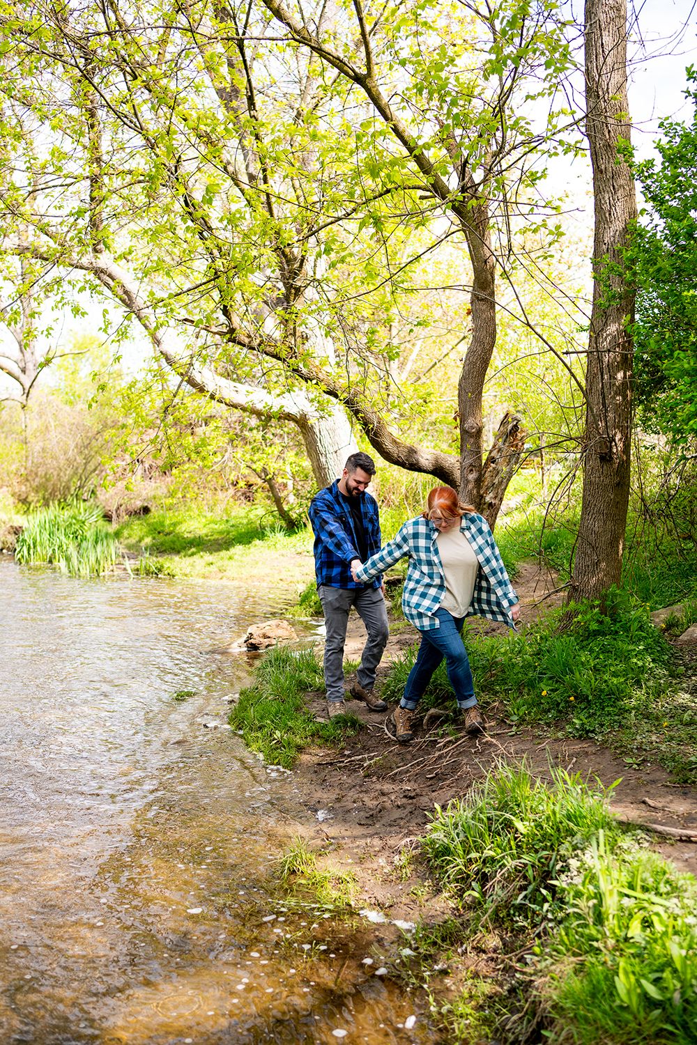 People explore along a wooded river bank with overhanging trees and flowing water in springtime.