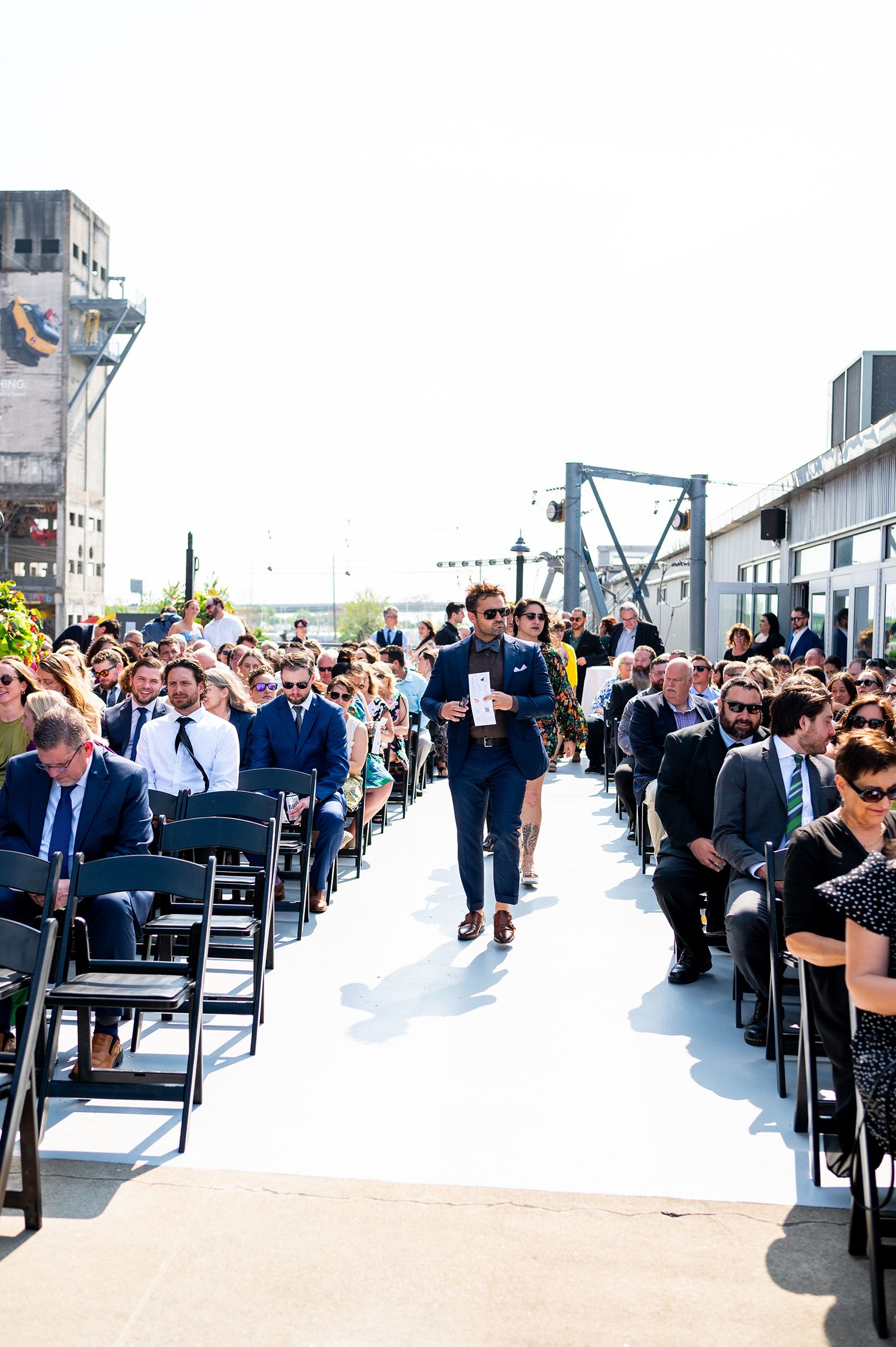 Wedding guests seated in rows of chairs on a white aisle runner at an outdoor industrial venue.