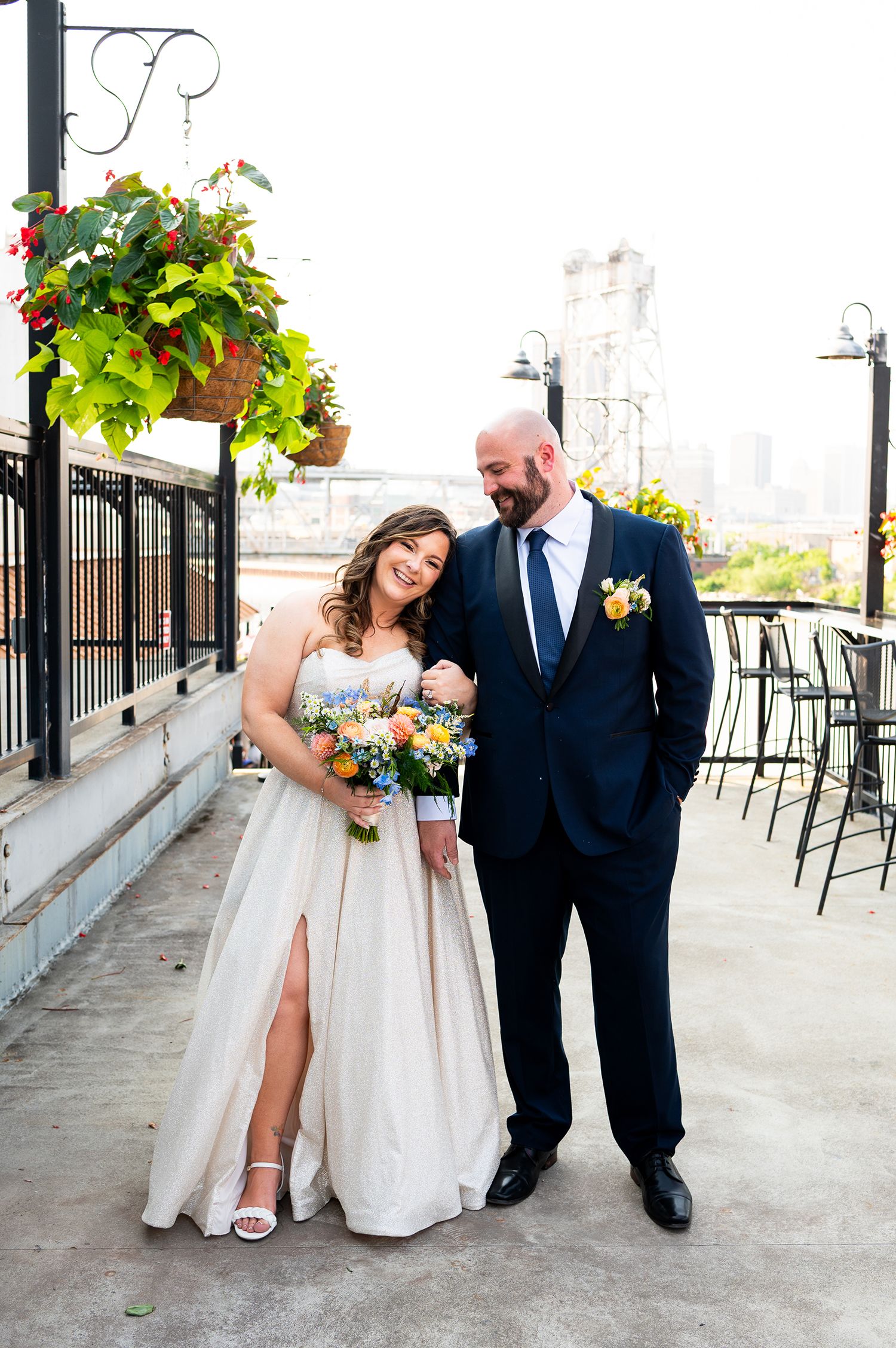 A couple in wedding attire poses along a stone wall with hanging plants and outdoor patio furniture.