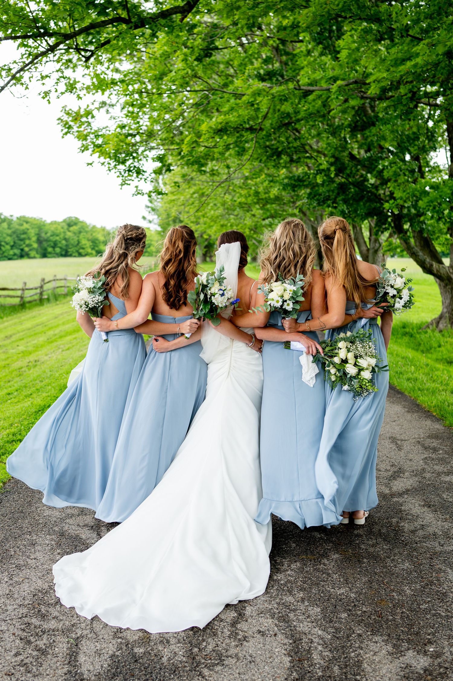 Wedding party in light blue bridesmaid dresses and white bridal gown pose together on a country path surrounded by greenery.