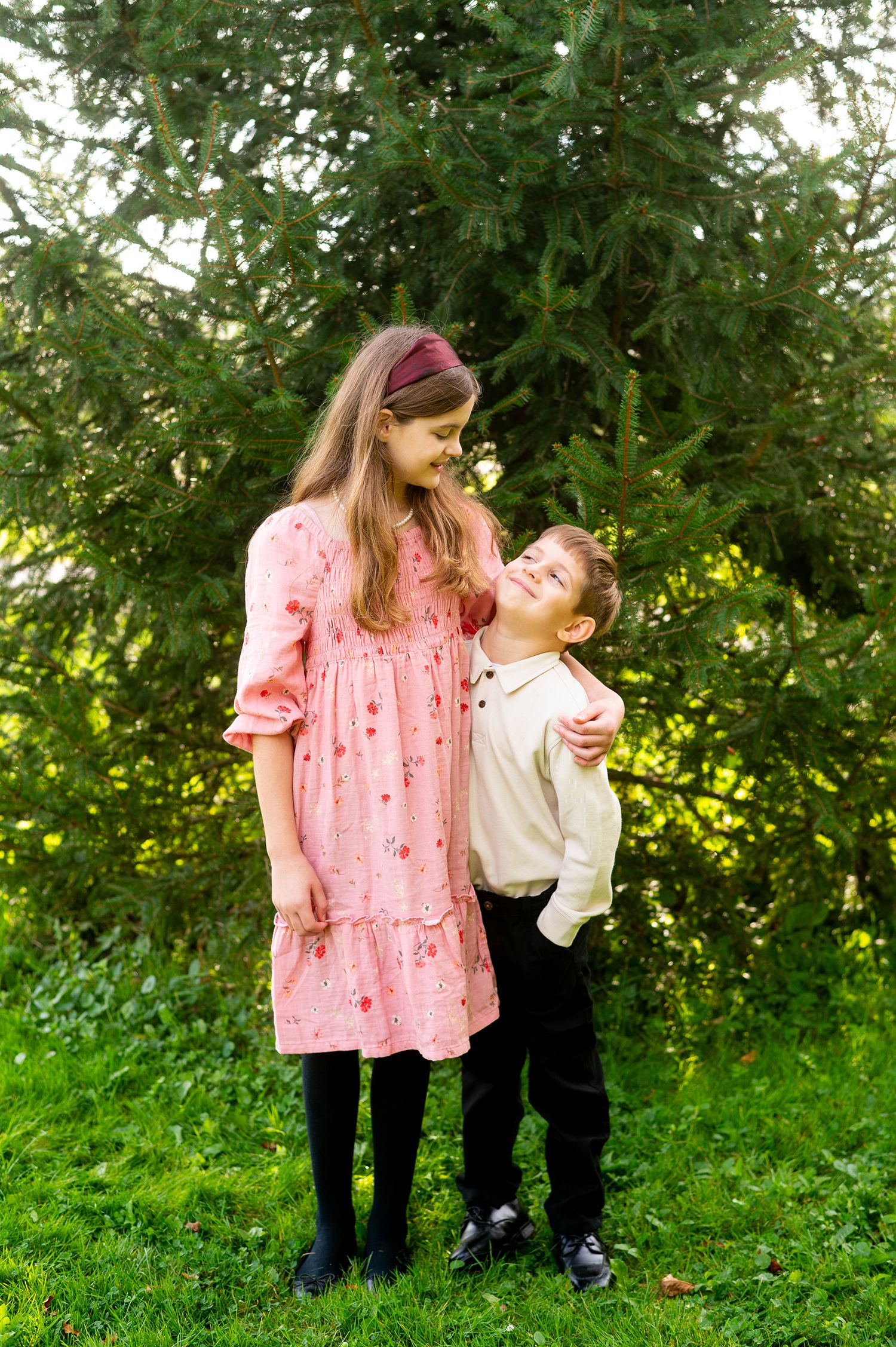 A girl in a pink dress and a boy in a white shirt share a warm embrace outdoors near evergreen trees.