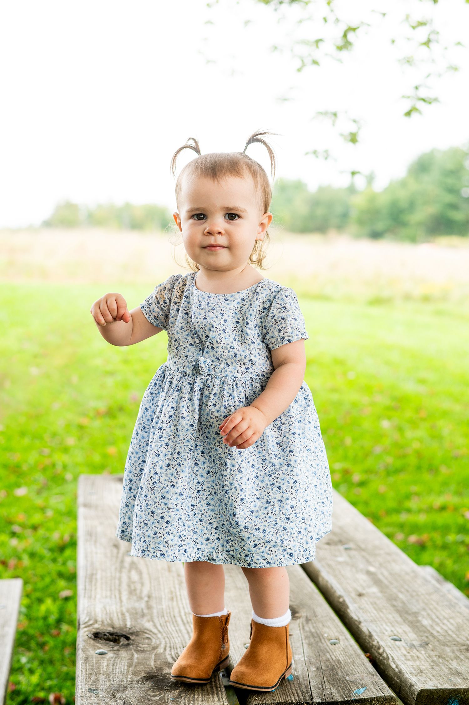 Adorable toddler girl in floral dress and brown boots standing on wooden bench in green outdoor field.