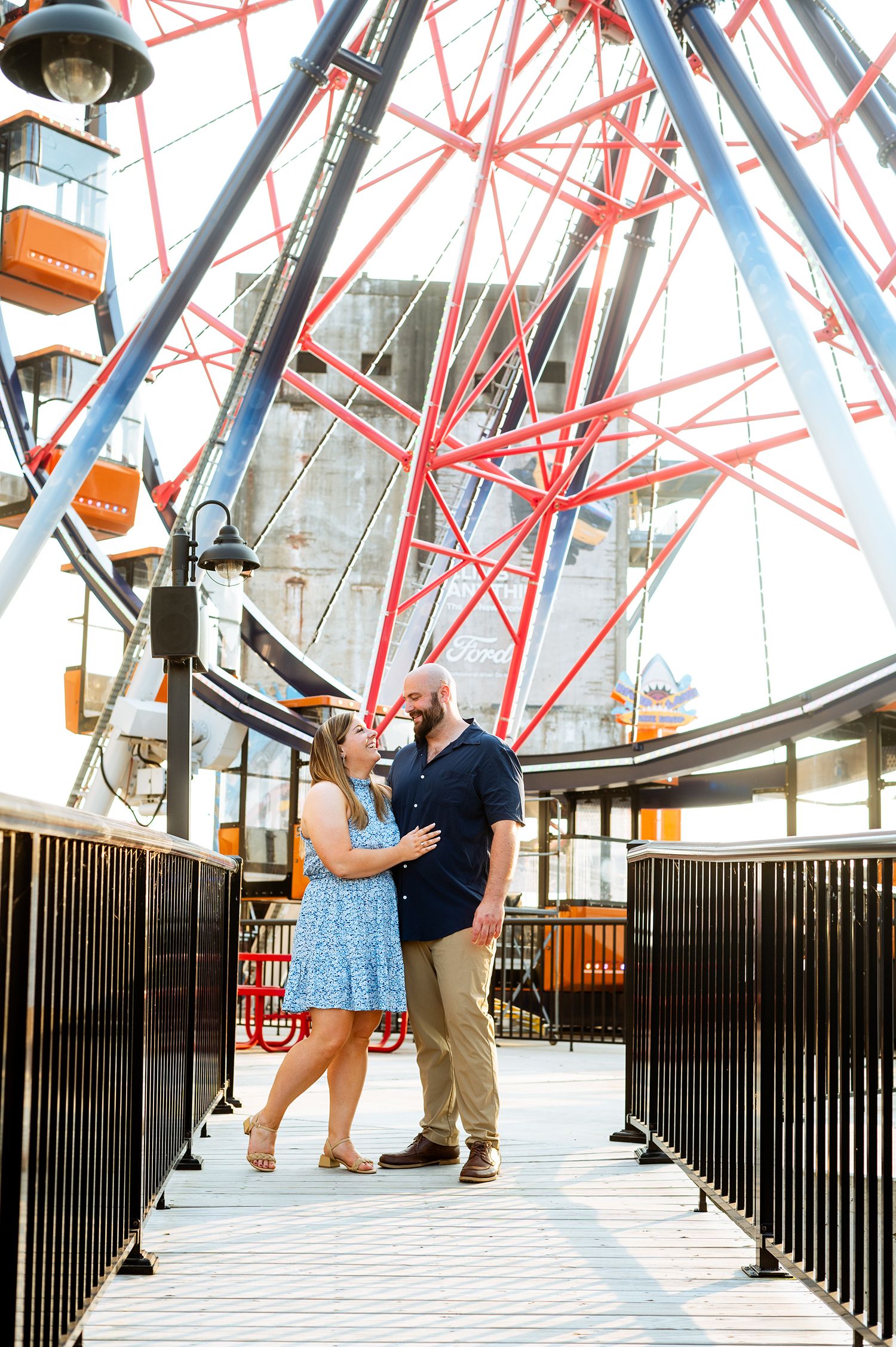 Couple embracing on a boardwalk in front of a colorful Ferris wheel during an outdoor engagement session.