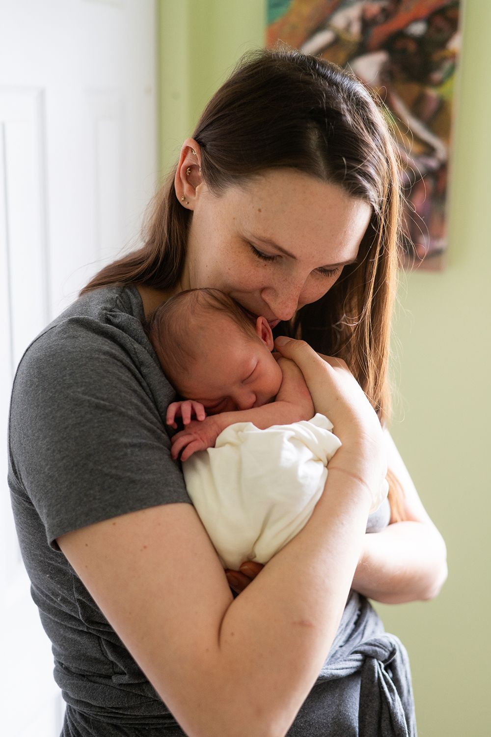 Parent in gray t-shirt tenderly kissing sleeping newborn baby wrapped in white blanket near window.