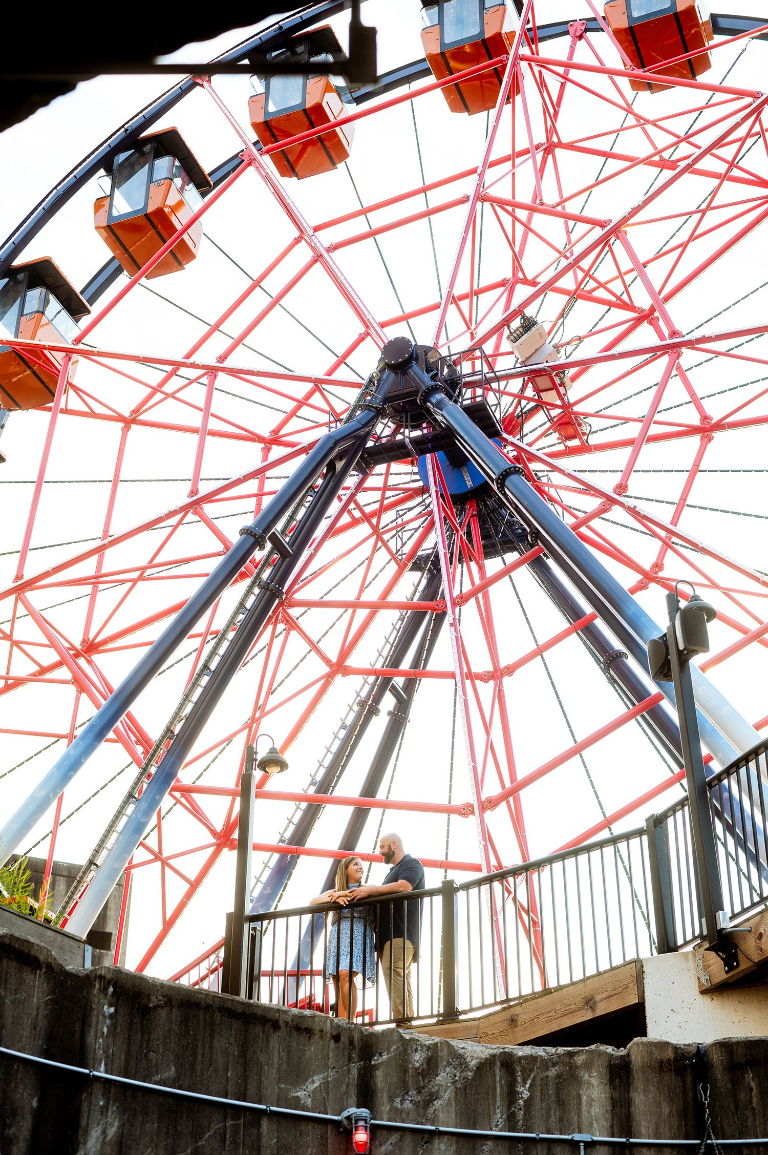 Two people stand beneath a large red and black Ferris wheel at an amusement park on a sunny day.