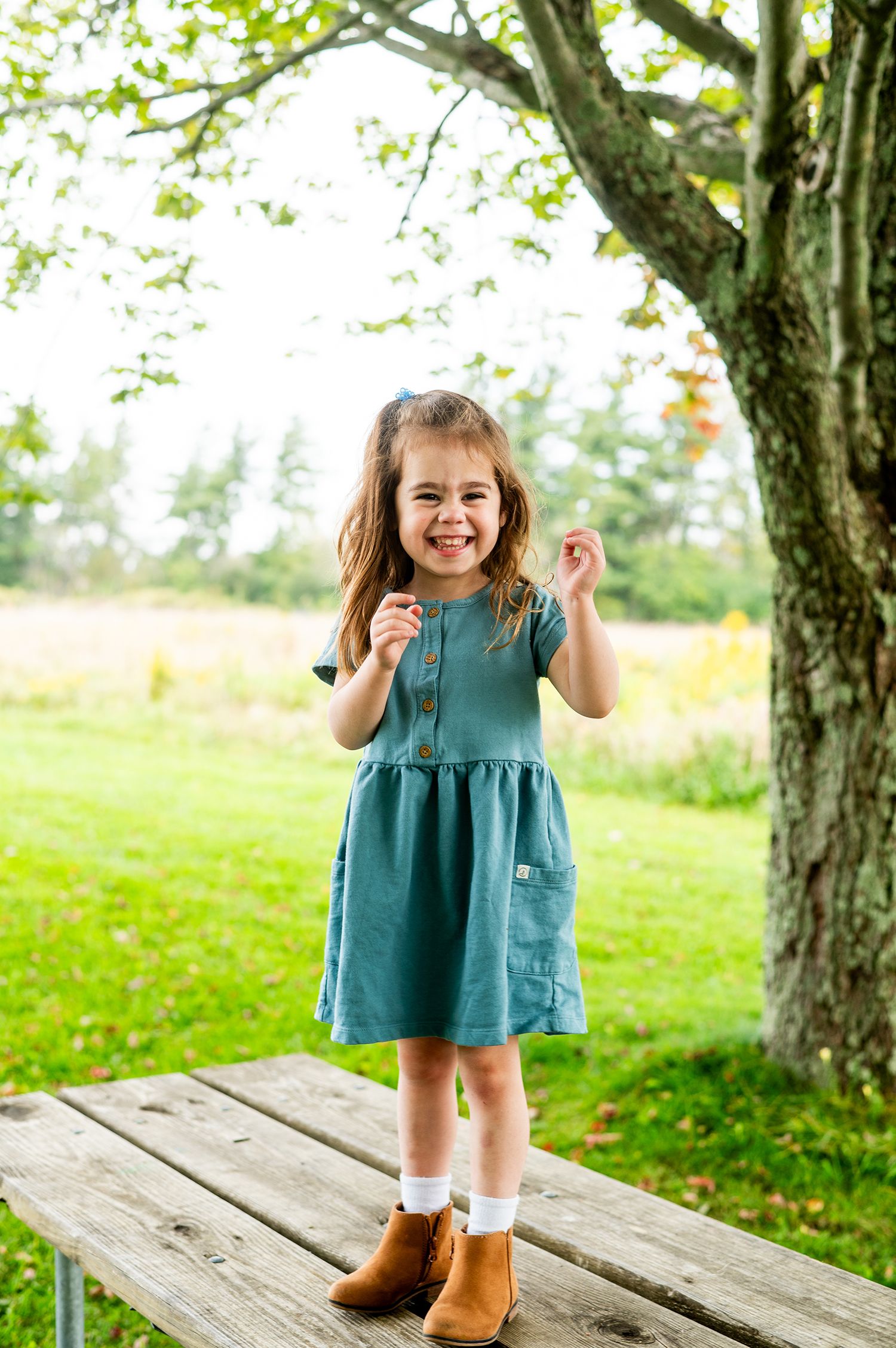 A cheerful young girl in a teal dress and brown boots stands on a wooden bench outdoors, smiling joyfully.