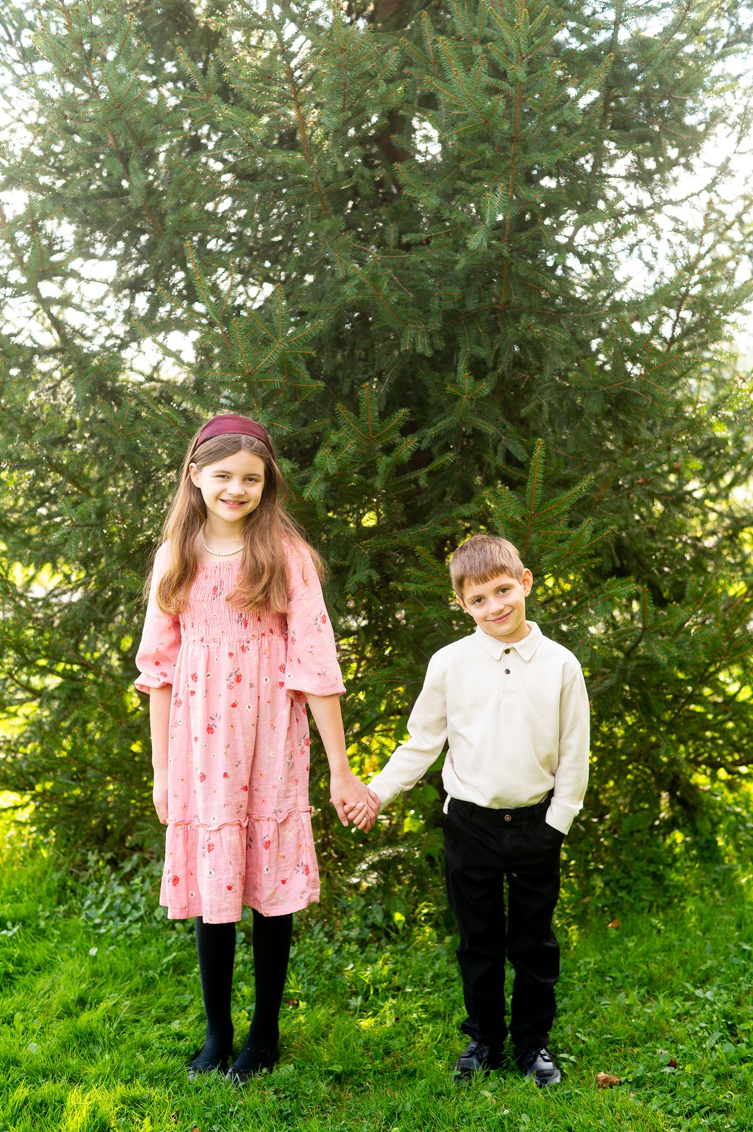 Two children, a girl in a pink dress and a boy in white shirt, hold hands smiling outdoors near evergreen trees.