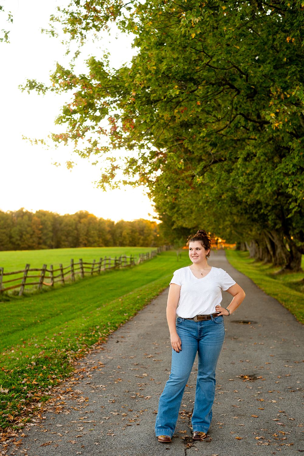 A tree-lined rural path with a wooden fence and green field at golden hour, featuring someone in casual attire.