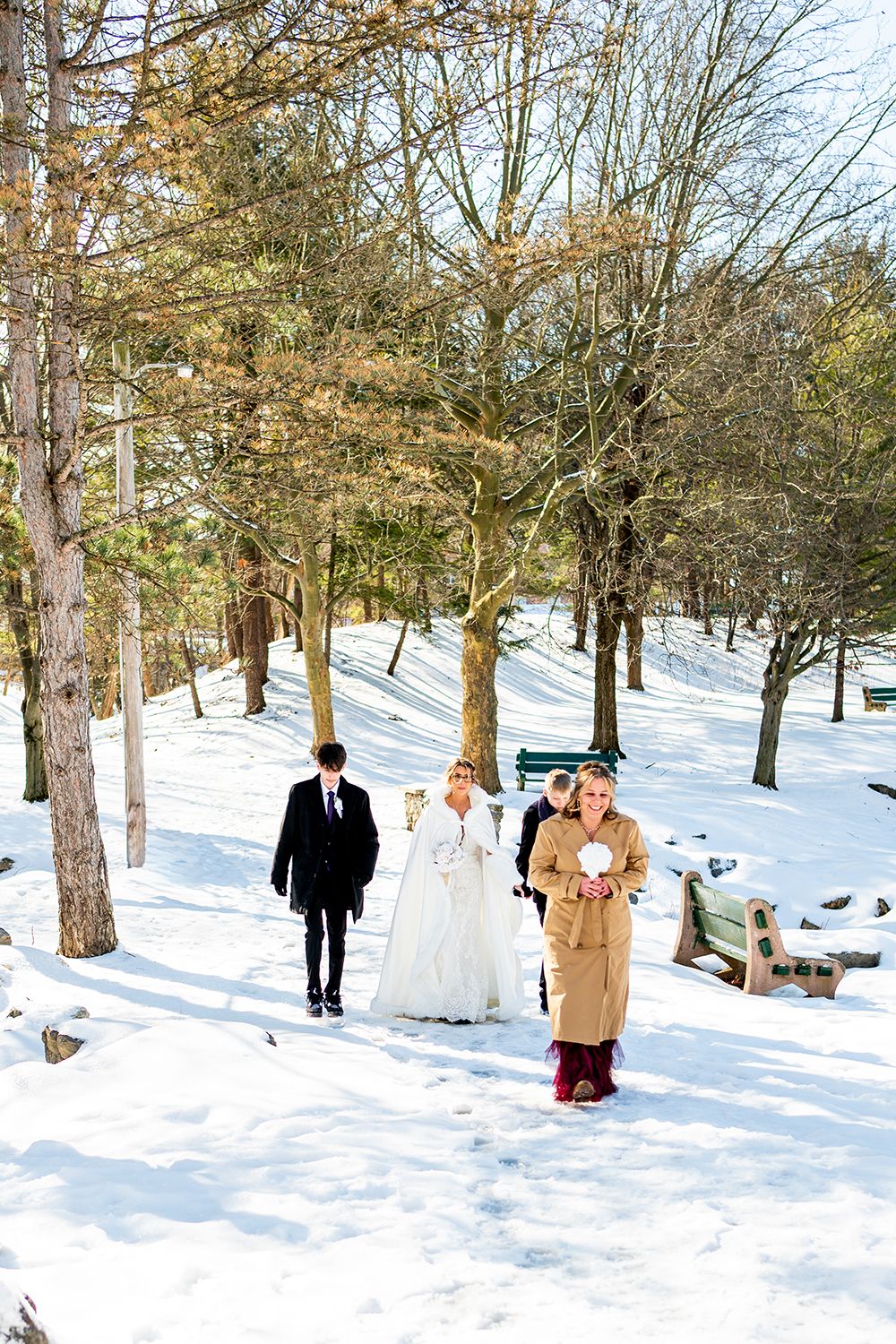 A winter wedding party walks through a snow-covered wooded path with bare trees and white ground.