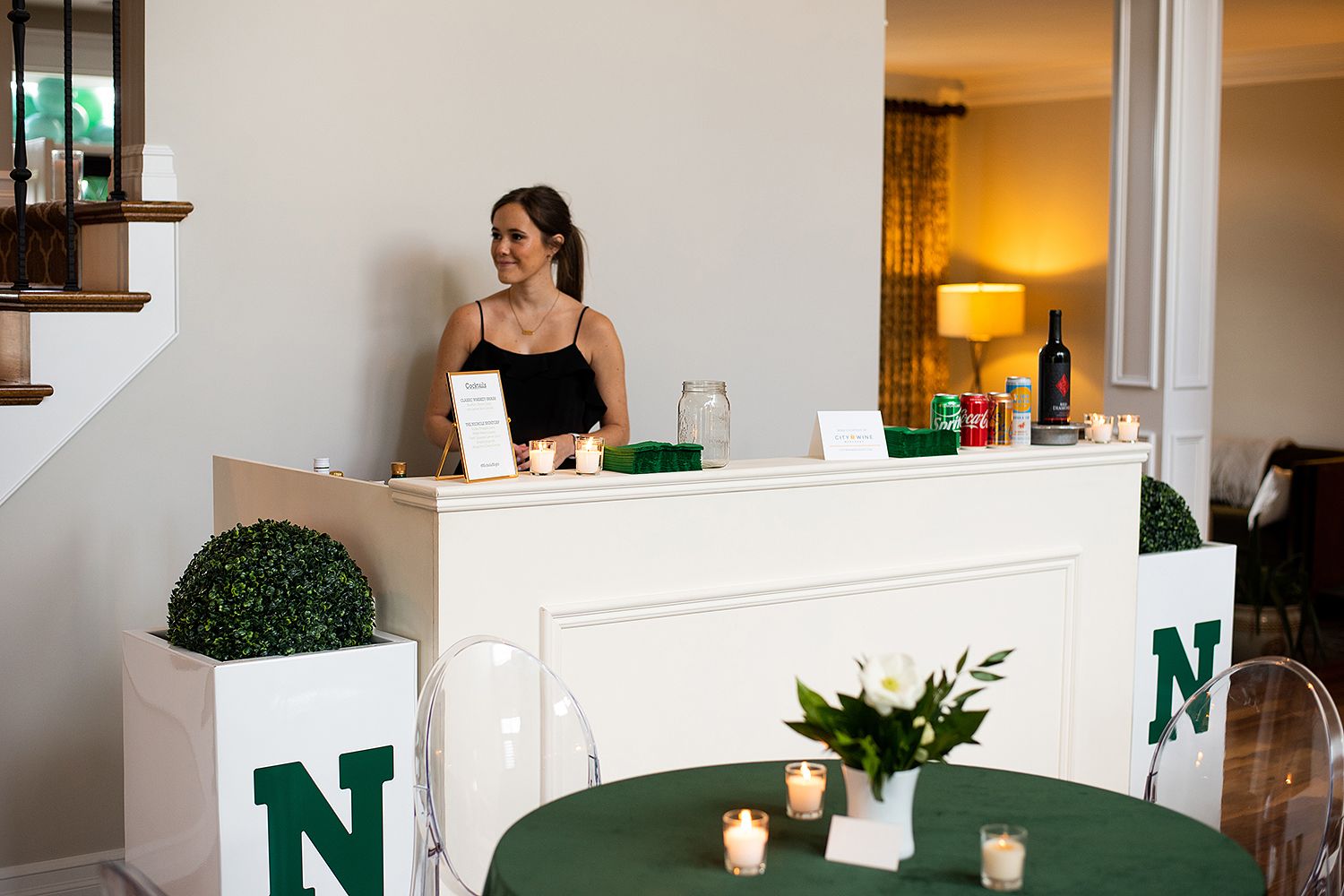 Someone tends to the white bar station decorated with green 'N' letters and boxwood plants at an upscale event.