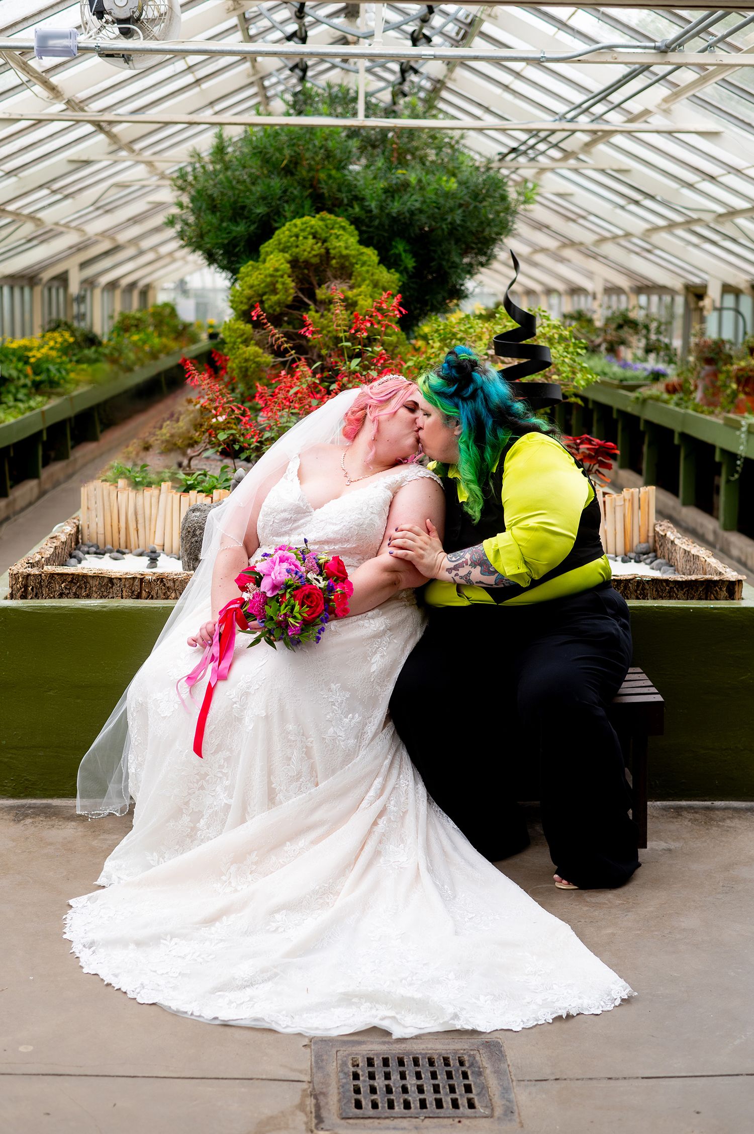 Two brides share a kiss in a greenhouse, one in a white lace gown with pink hair, the other in a green jacket with teal hair.