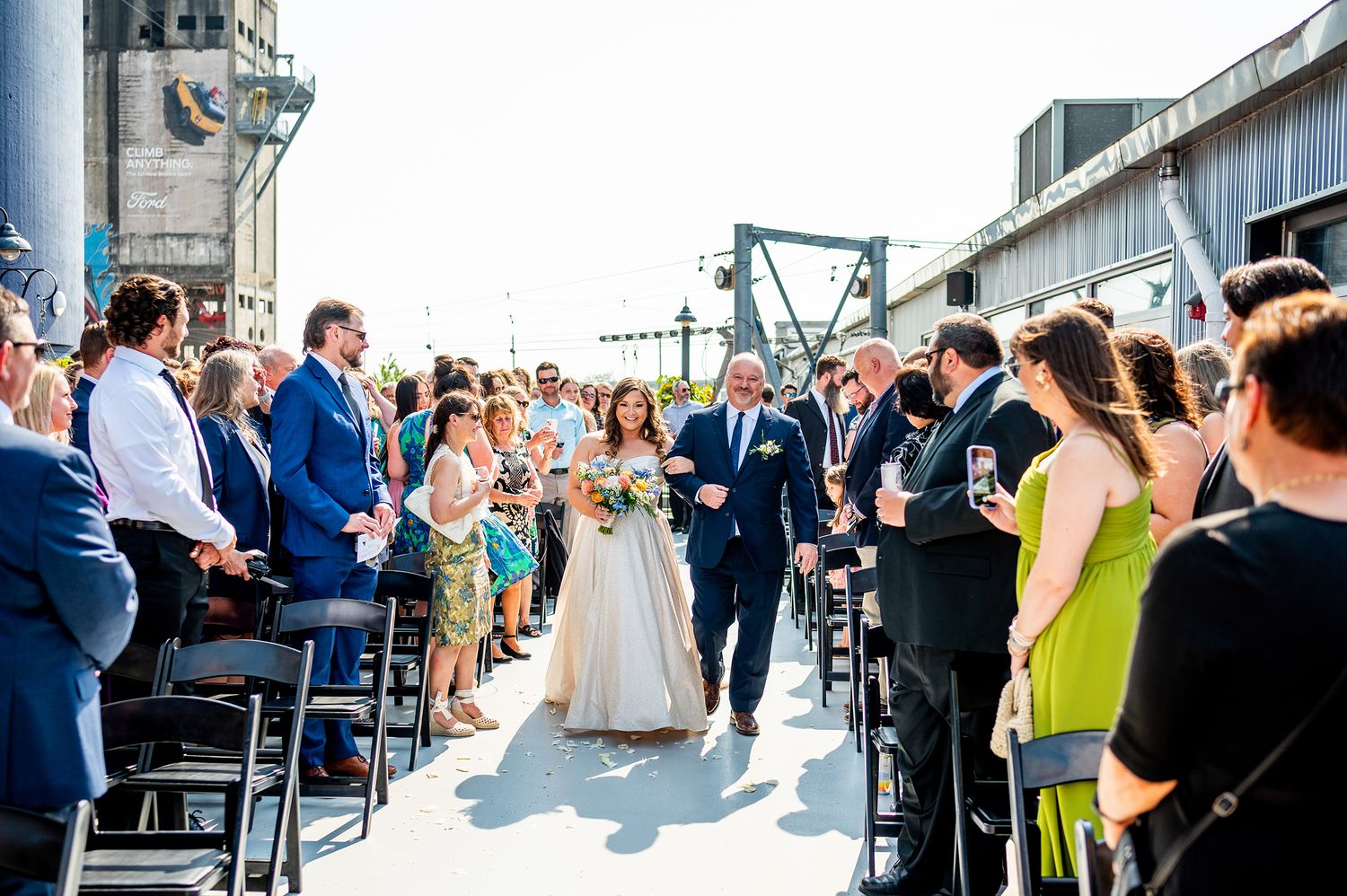 Wedding guests gather outside a rustic barn venue on a sunny day to celebrate the newlyweds.