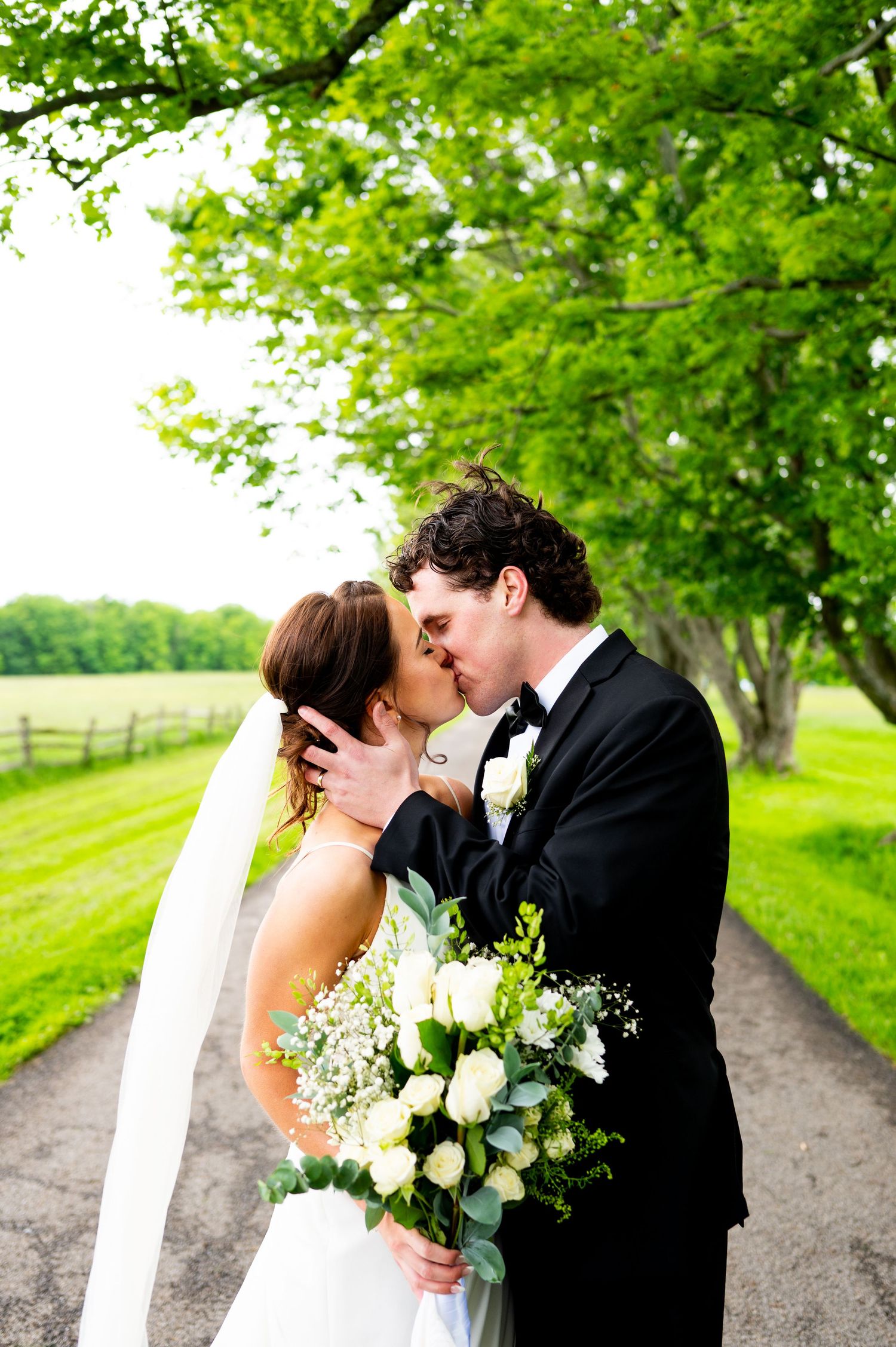 A romantic wedding couple shares a tender kiss on a tree-lined path surrounded by lush green foliage.