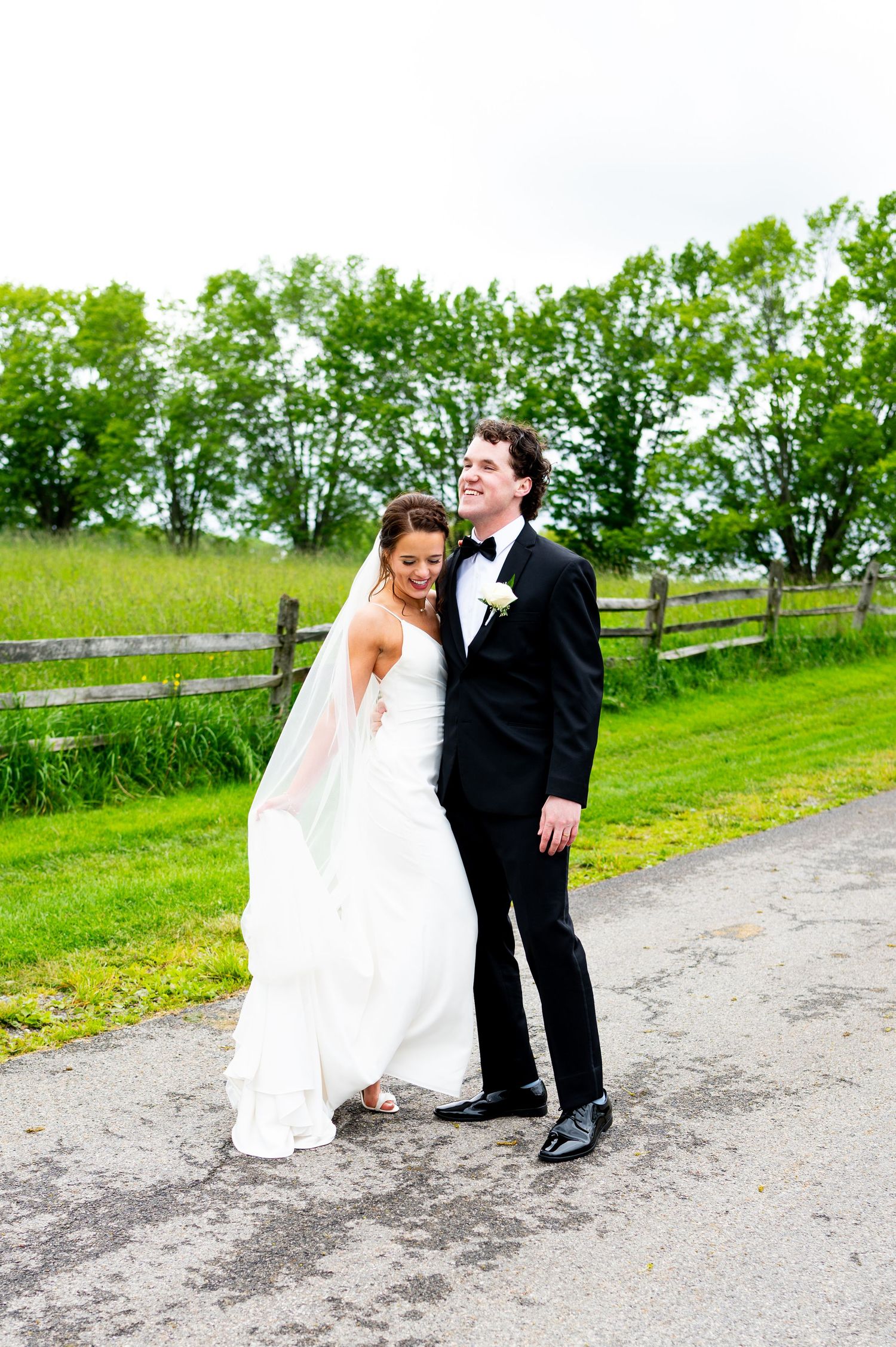 A couple in wedding attire share a romantic moment on a country dirt road with green fields and wooden fence.