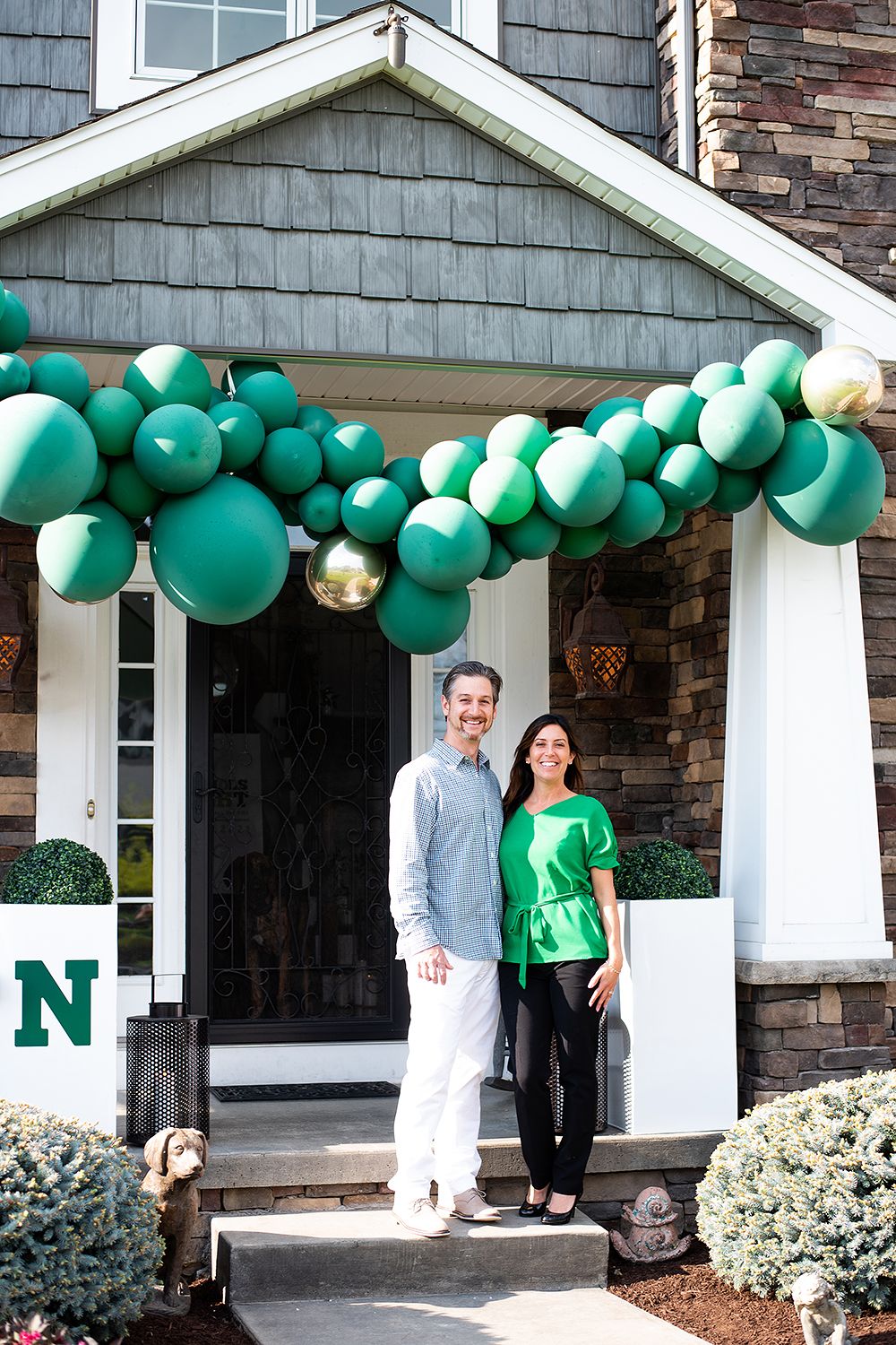 A home entrance decorated with a festive arch of green and gold balloons along the front porch columns.