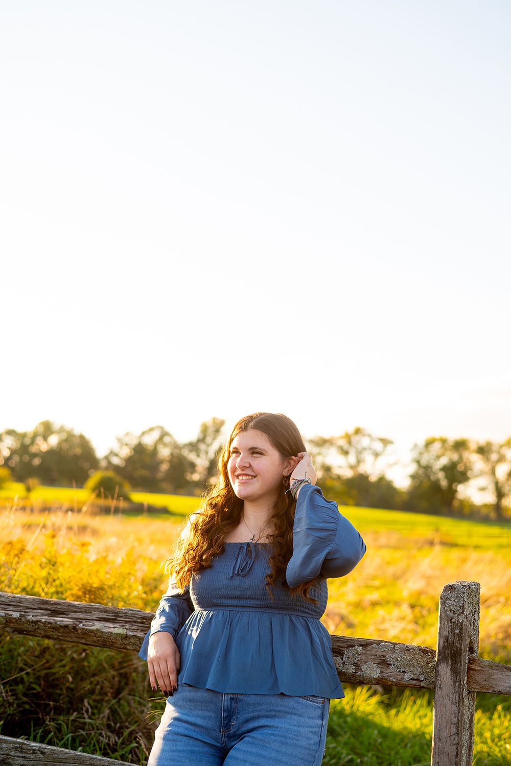 Person in denim outfit leans against rustic fence during golden hour in a countryside meadow setting.